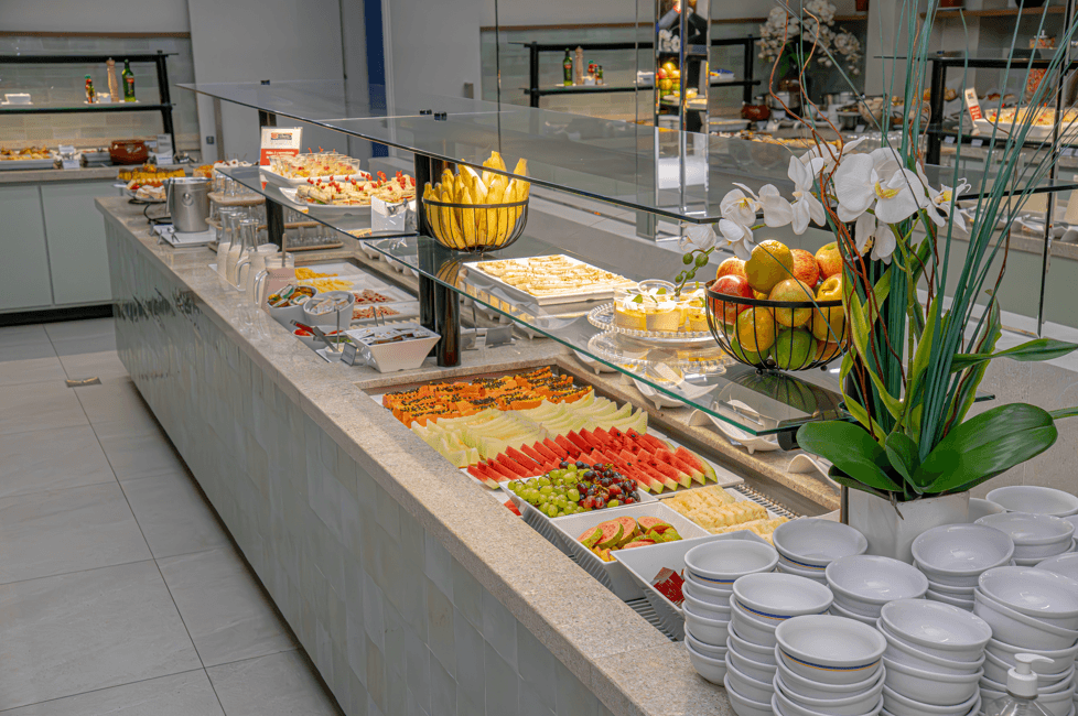 Mesa de bufê com comida. Frutas, doces e acompanhamentos expostos; tigelas brancas empilhadas.