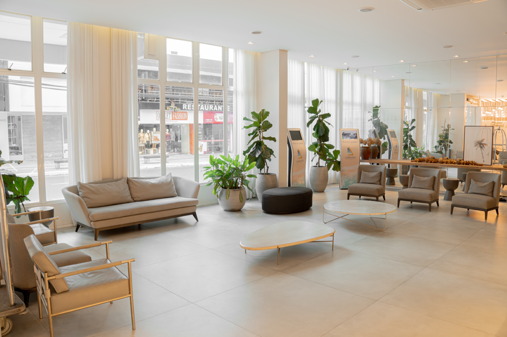 Saguão iluminado e arejado com sofás, poltronas, plantas e grandes janelas com vista para uma cena de rua.