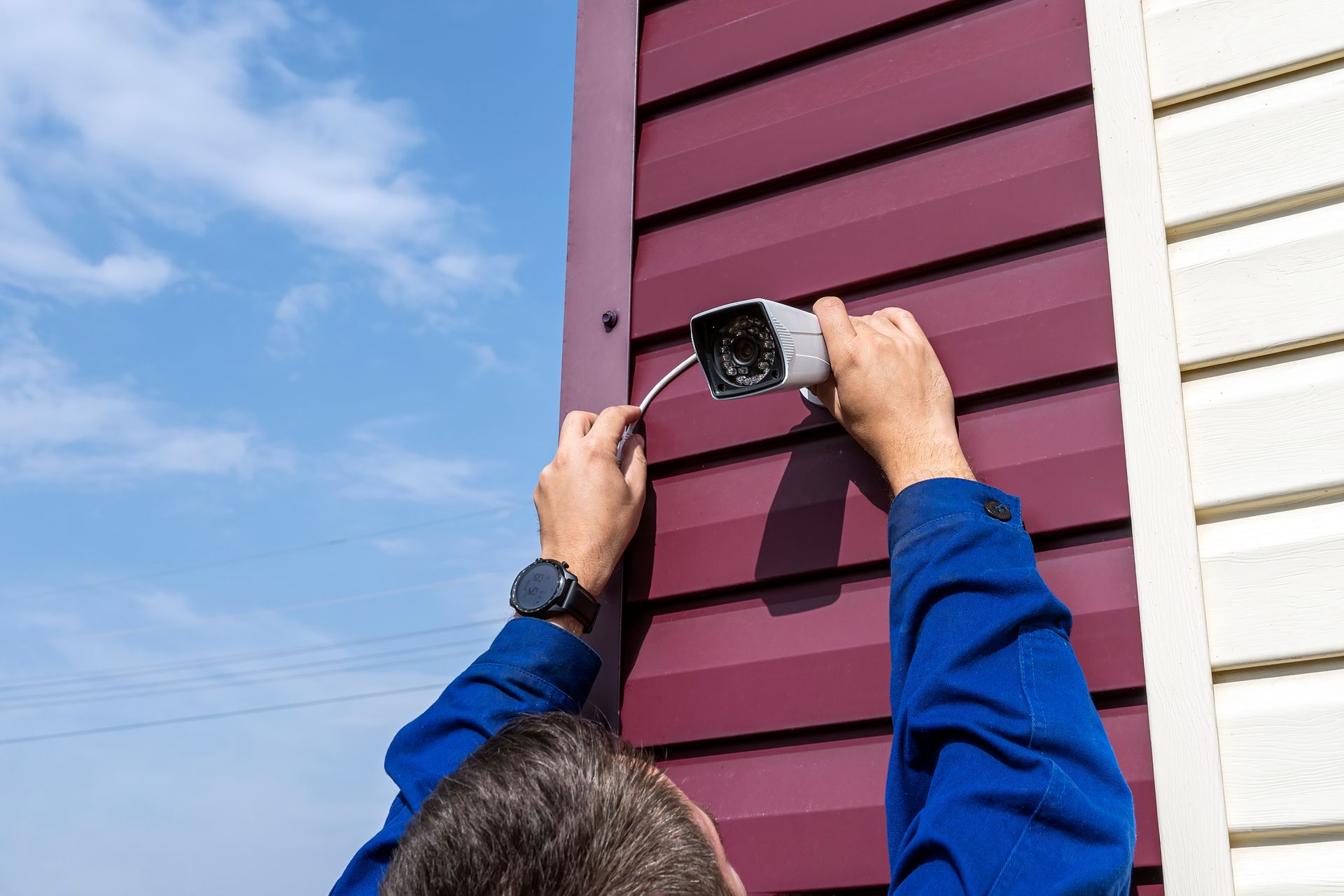 Person installing a security camera on a burgundy siding wall under a blue sky. Person installing a security camera on a burgundy siding wall under a blue sky.