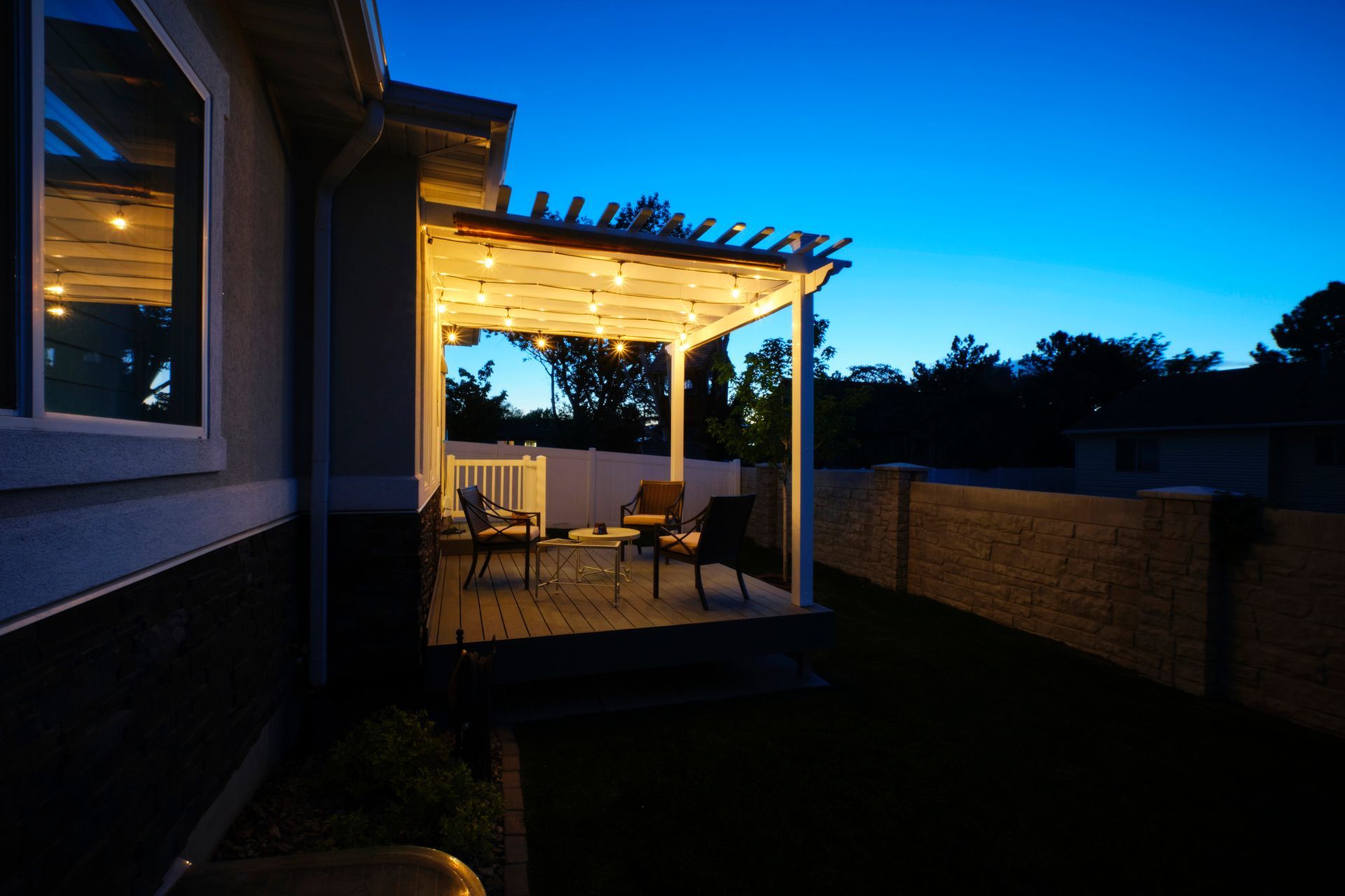 A patio with a pergola and chairs is lit up at night.
