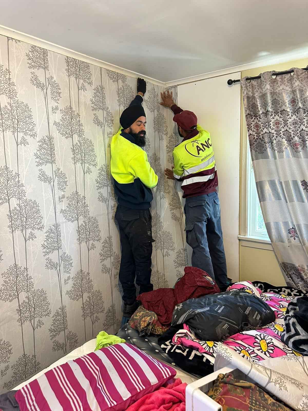 Two men are hanging wallpaper on a wall in a bedroom.
