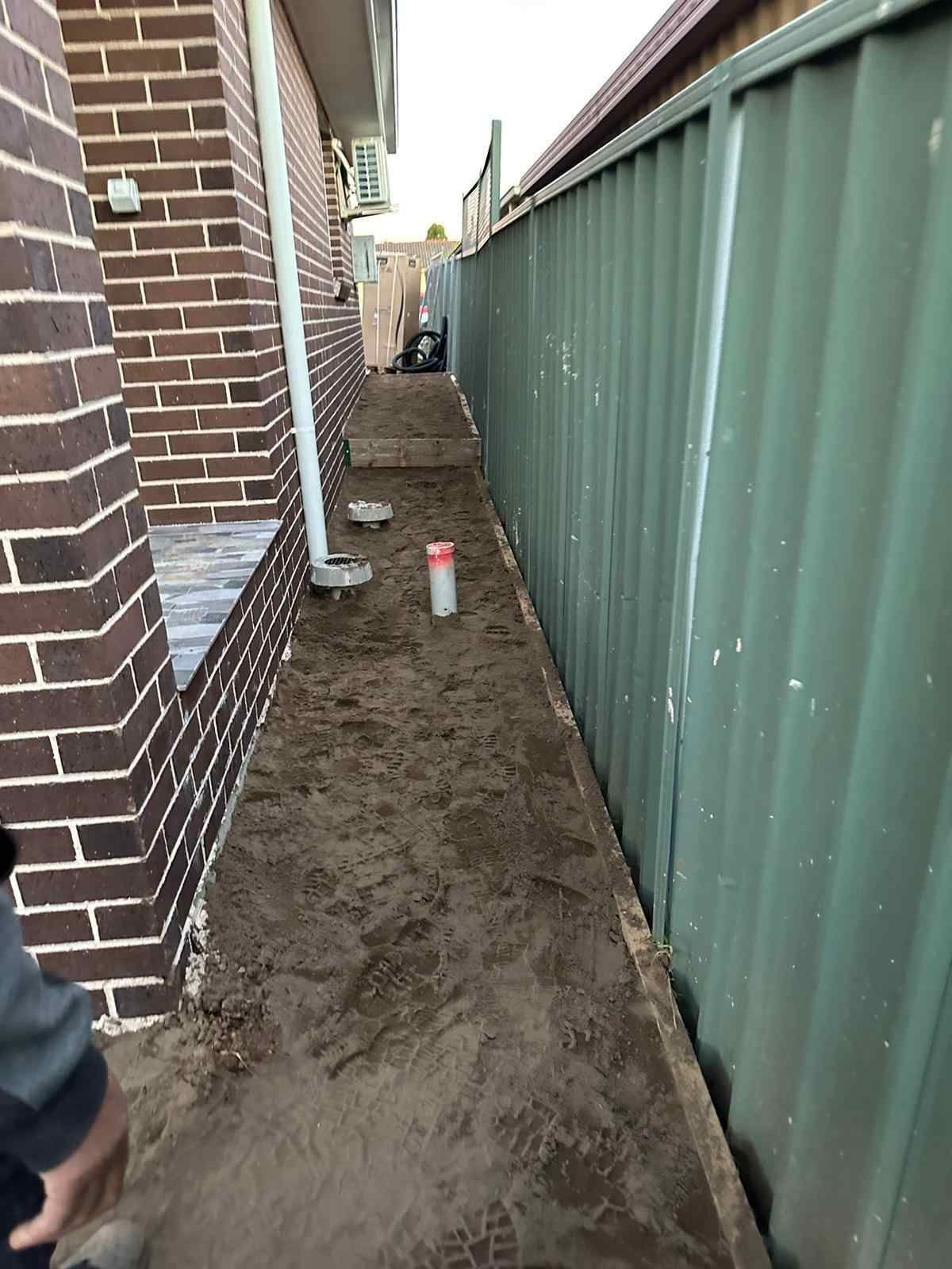 A person is standing on a dirt path next to a green fence.