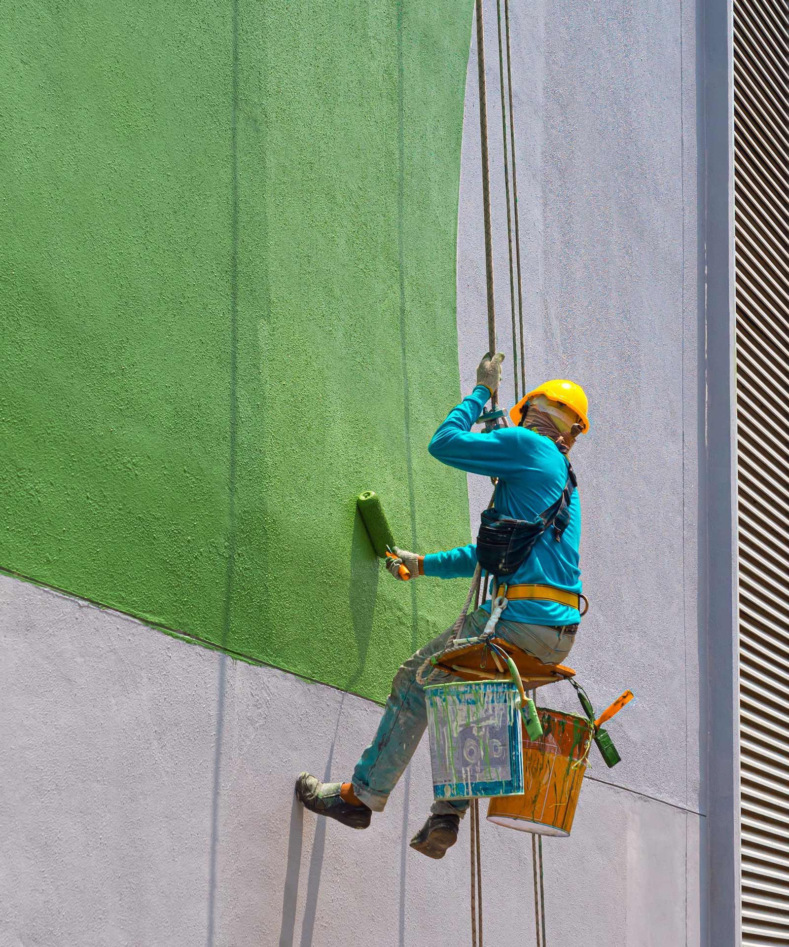 A man is painting a building with a roller while hanging from a rope.