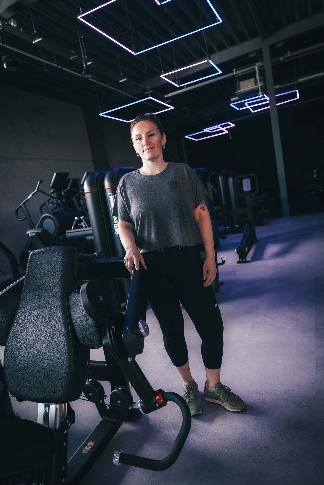 Een vrouw staat bij een fitnessapparaat in de sportschool, gekleed in een grijs shirt en een zwarte broek. Er hangen blauwe en witte lampen aan het plafond.