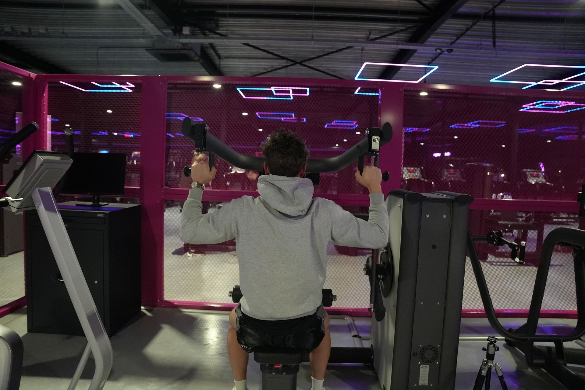 Een persoon gebruikt een lat pulldown-apparaat in een sportschool. Felroze en paarse lichten op de achtergrond.