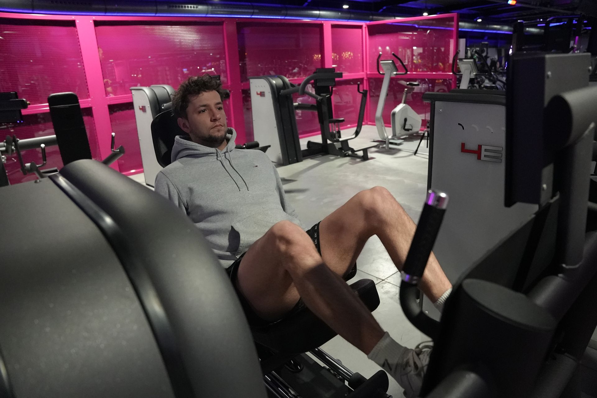Man in een grijze hoodie gebruikt een leg press-apparaat in een sportschool met roze muren.