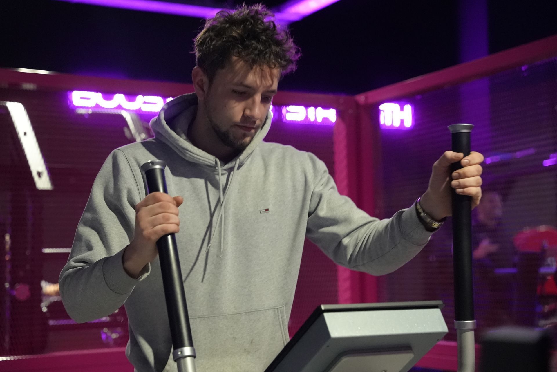 Man in grijze hoodie gebruikt een crosstrainer in een sportschool met paarse neonreclames.