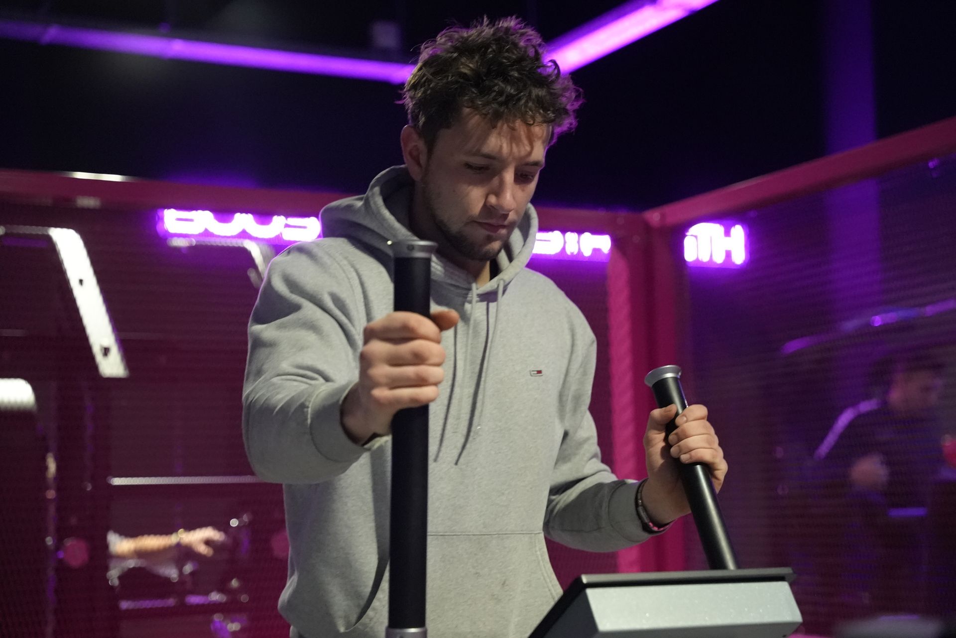 Man in grijze hoodie gebruikt een fitnessapparaat met zwarte handgrepen in een sportschool met paarse verlichting.