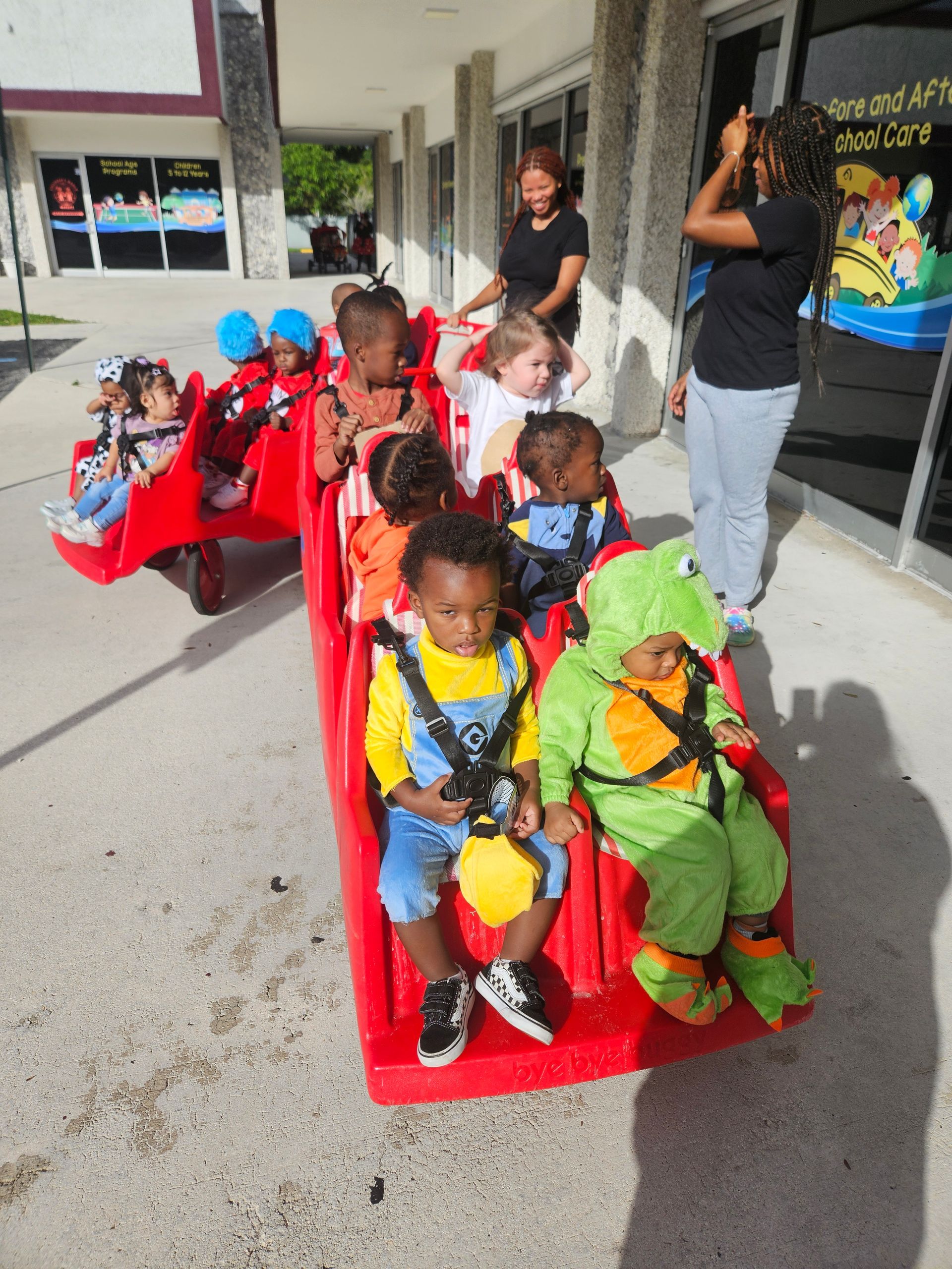 Children in costume sit in a red wagon, guided by two adults outside a building.