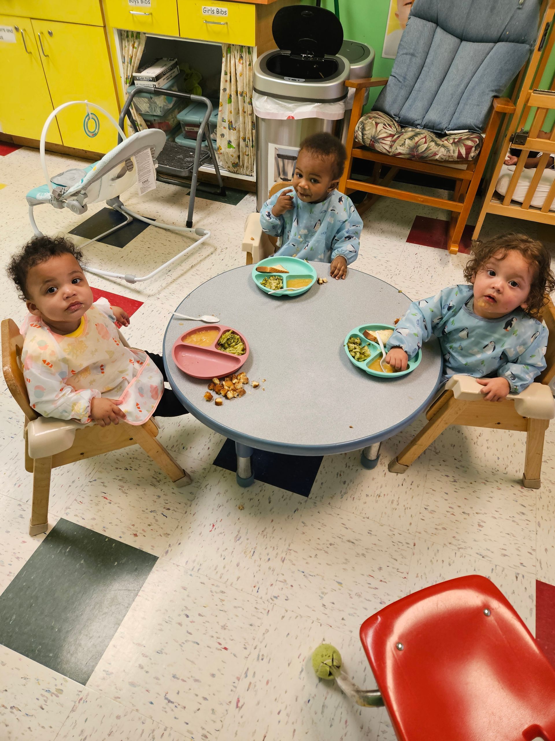Three babies eating at a round table in a classroom.