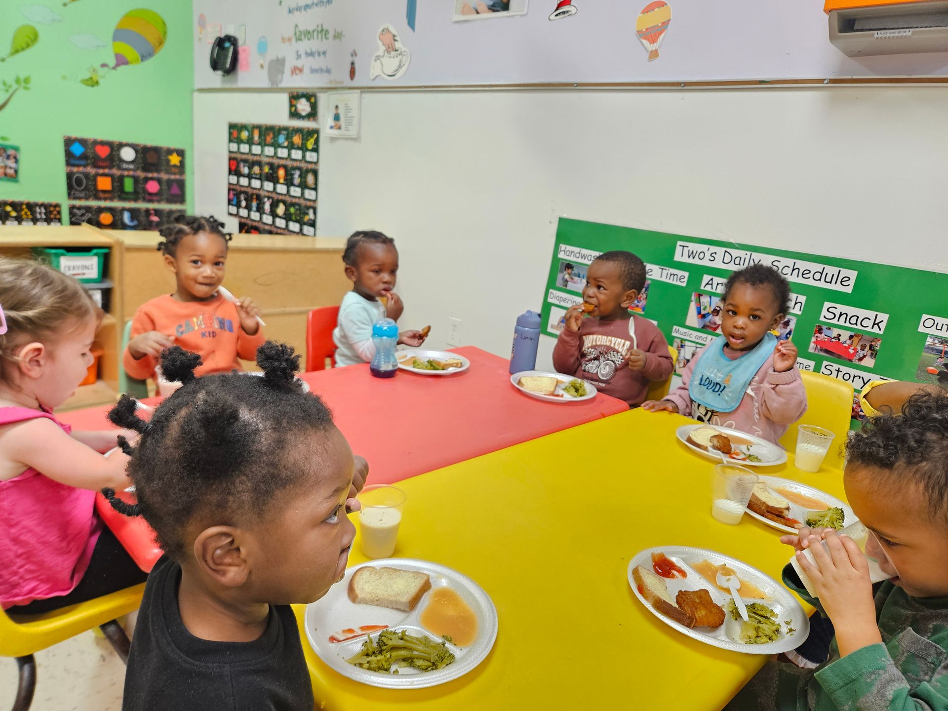 Children eating lunch at a table in a colorful classroom; some smiling, some eating.