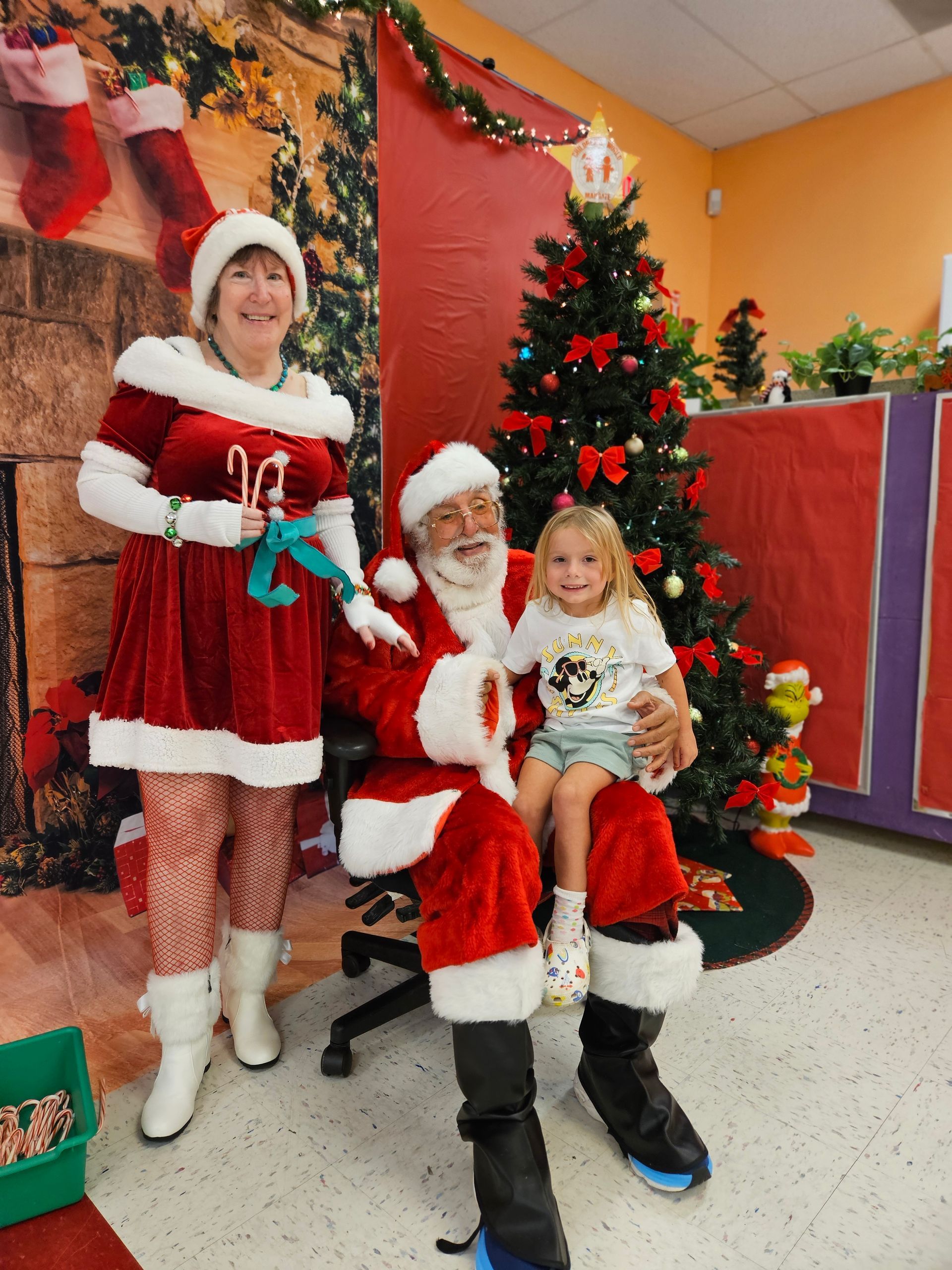 Woman in Santa outfit and Santa with a child at a Christmas scene.
