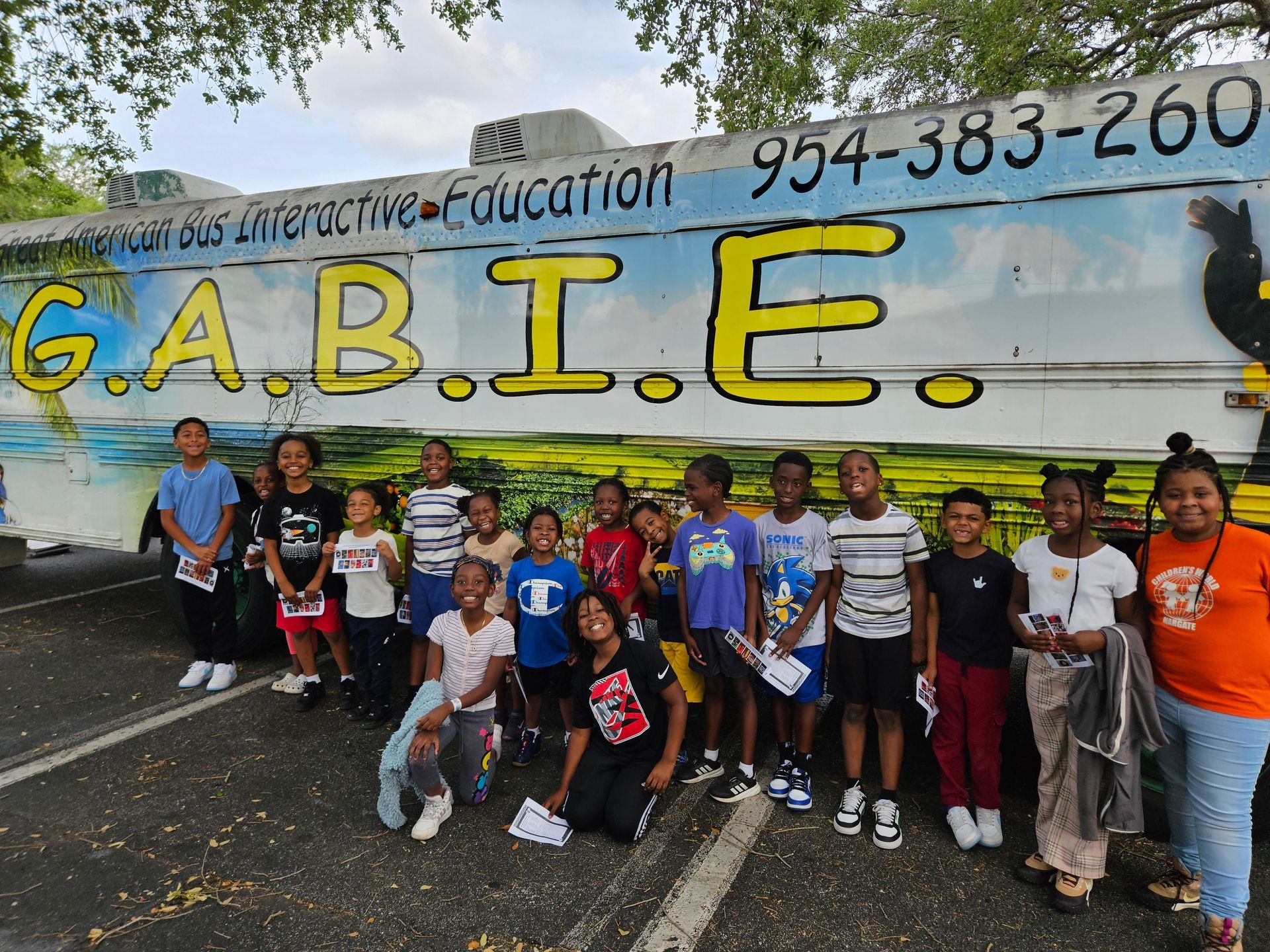 Group of kids posing in front of a colorful bus that reads G.A.B.I.E. outdoors on a sunny day.