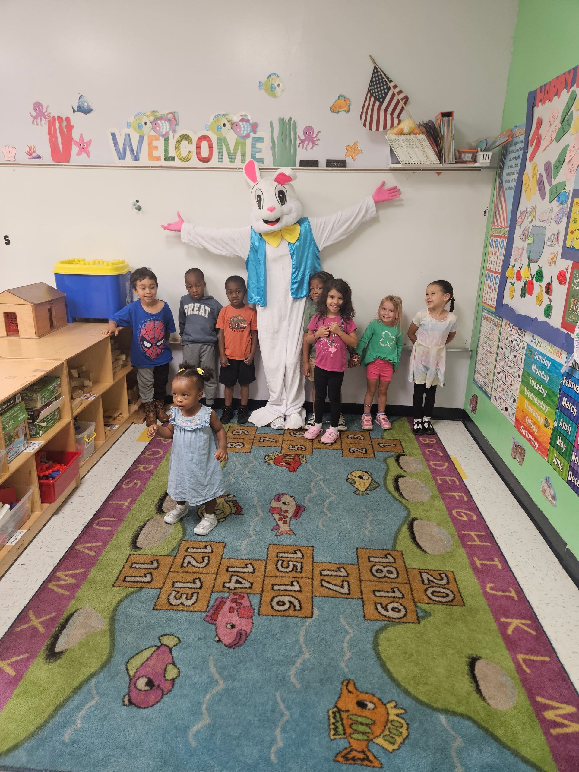 Children with Easter Bunny in a classroom; children, bunny are smiling, standing in front of a colorful rug and decorations.