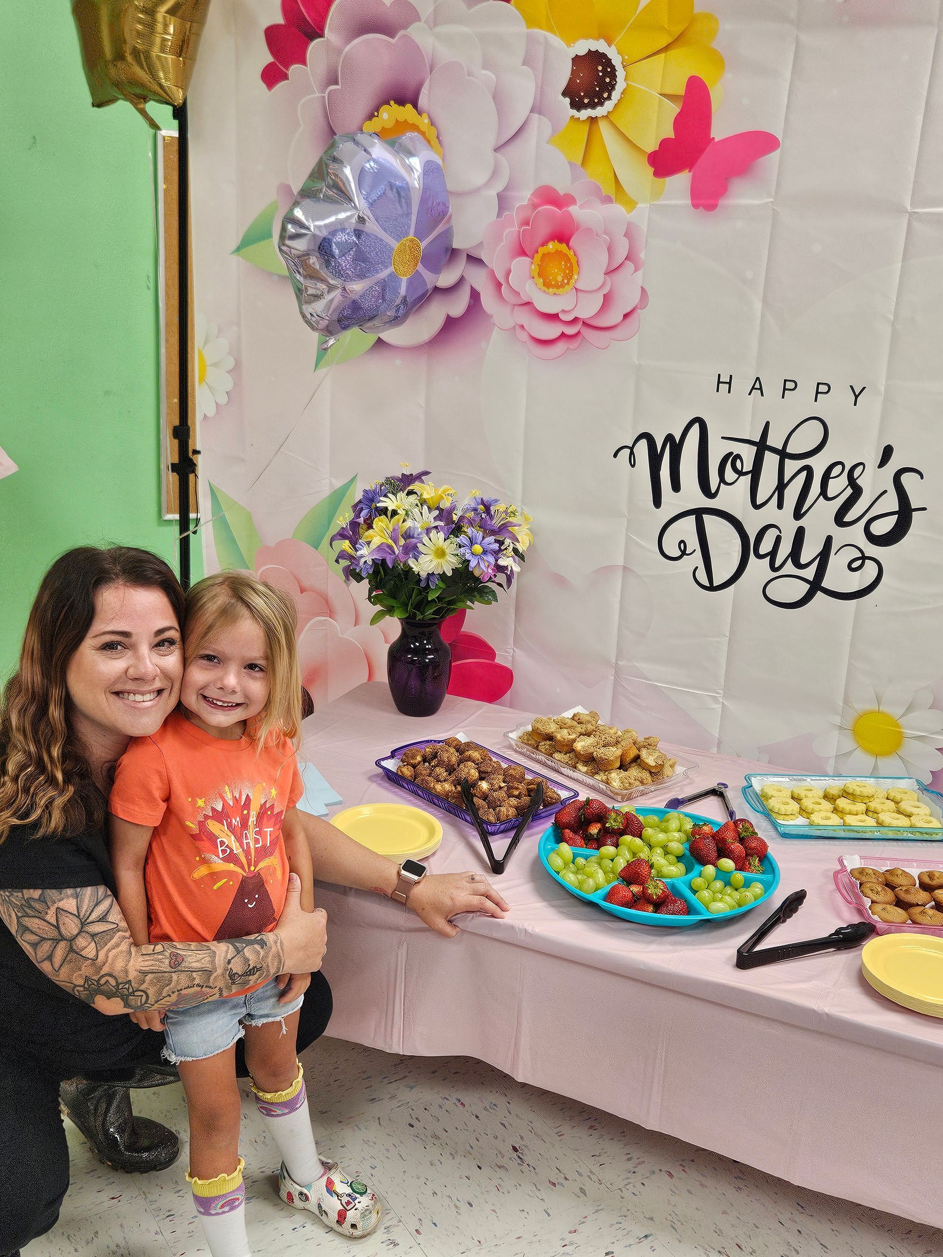 Woman and child at a Mother's Day party. They smile next to a table of food and floral decorations.