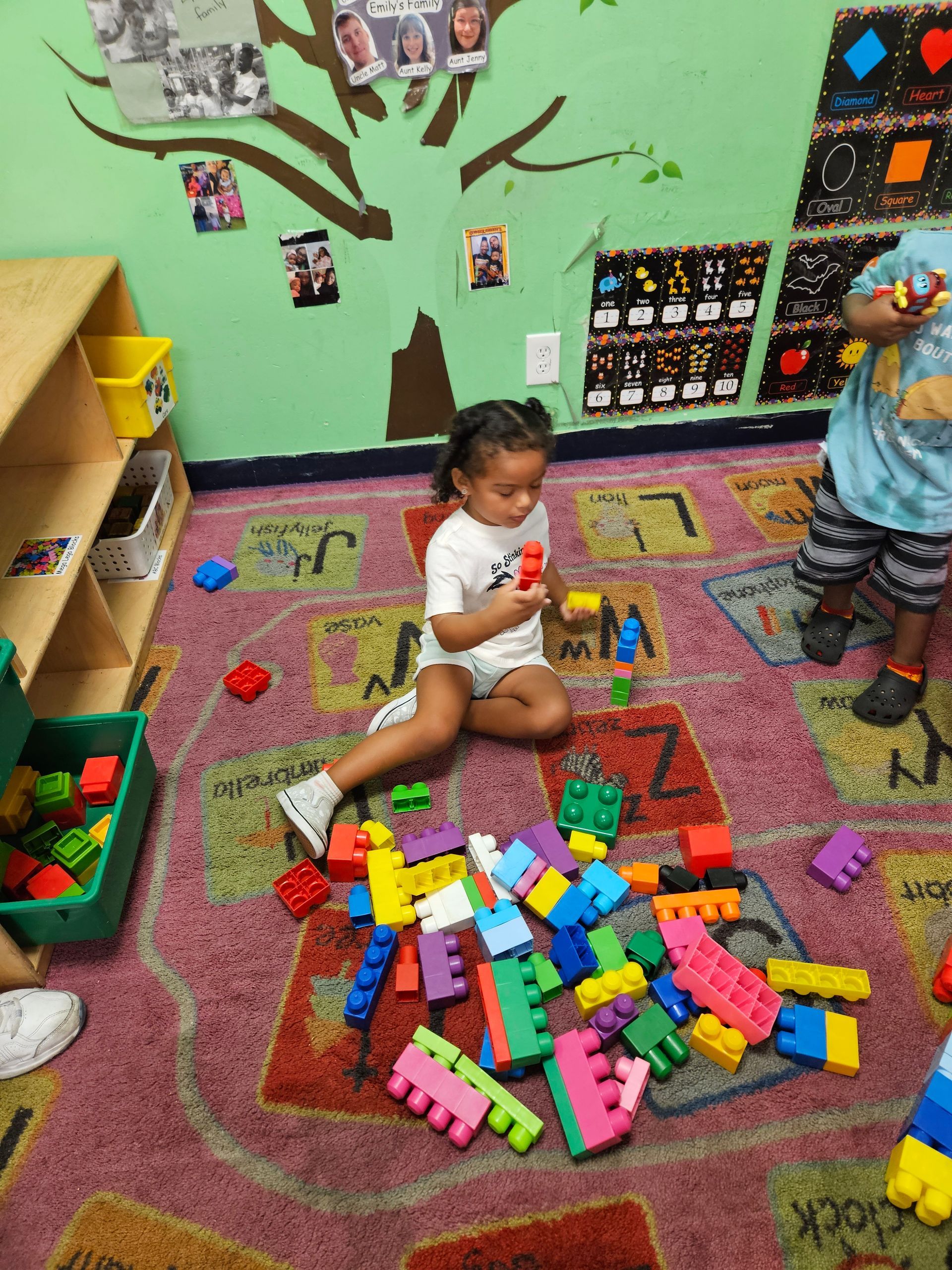Child playing with colorful blocks on a patterned rug in a classroom, another child nearby.