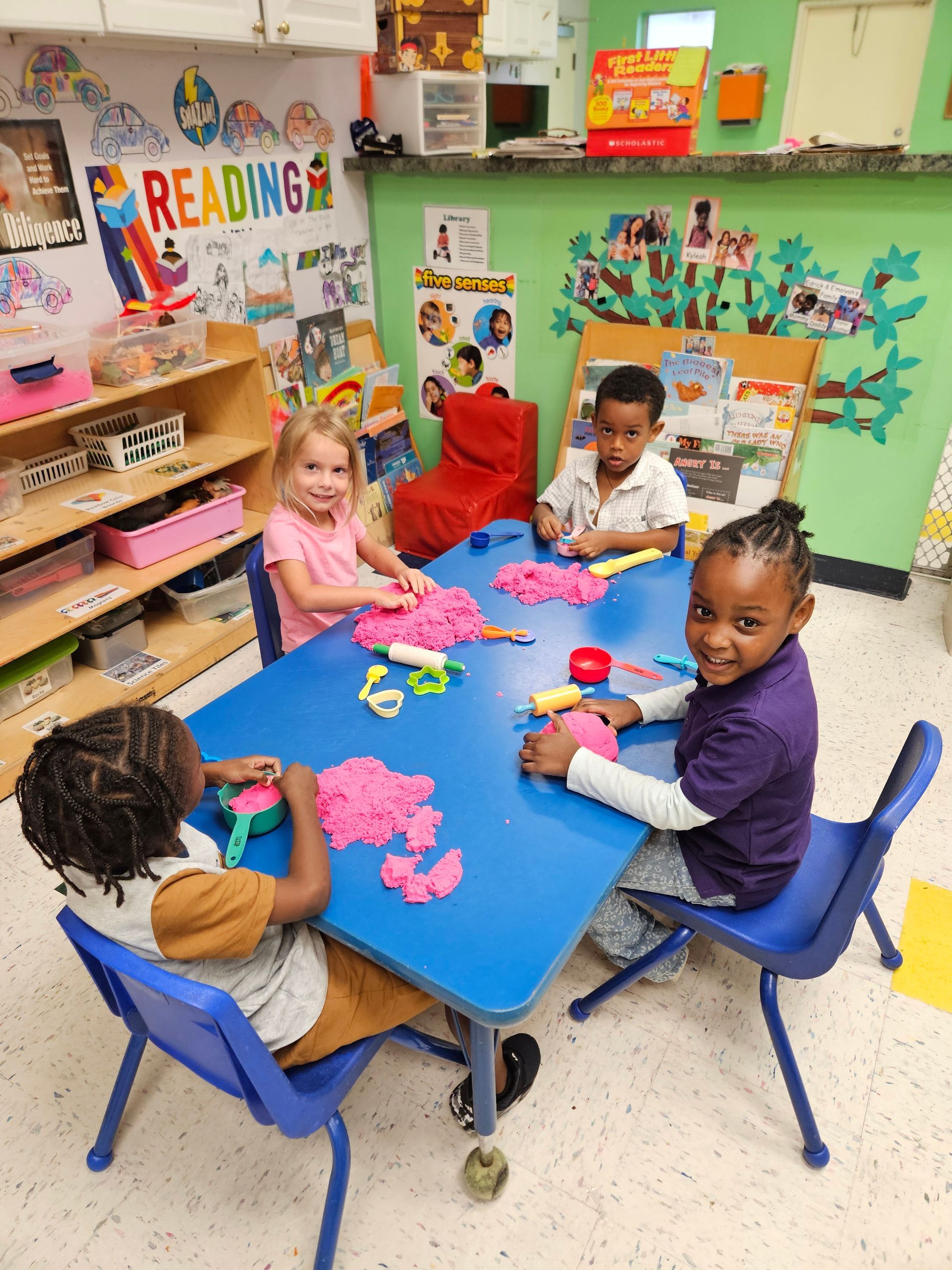 Four children at a blue table, playing with pink sensory sand in a brightly lit classroom.