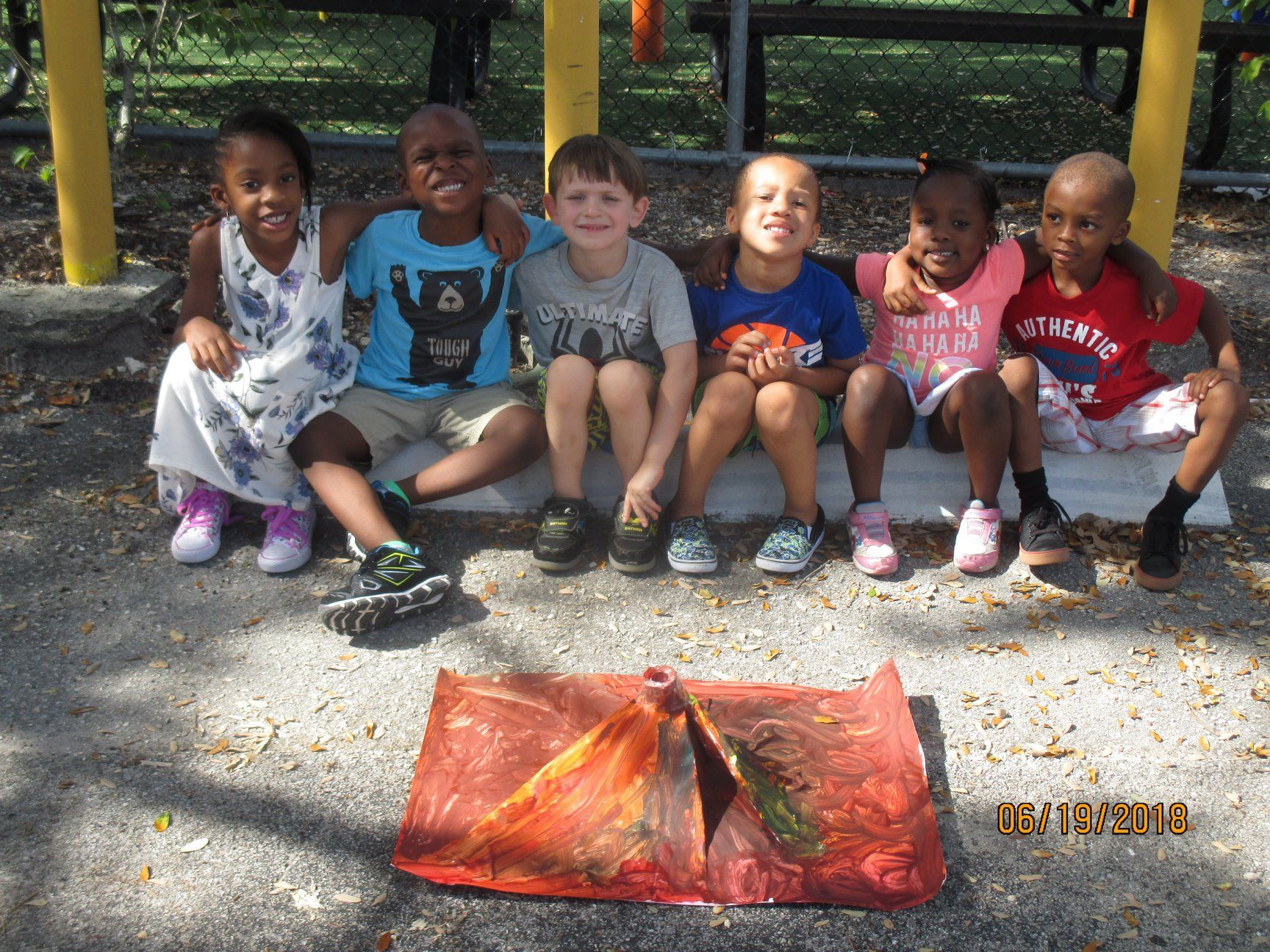 Six children smiling outdoors, sitting with arms around each other, in front of a volcano craft.