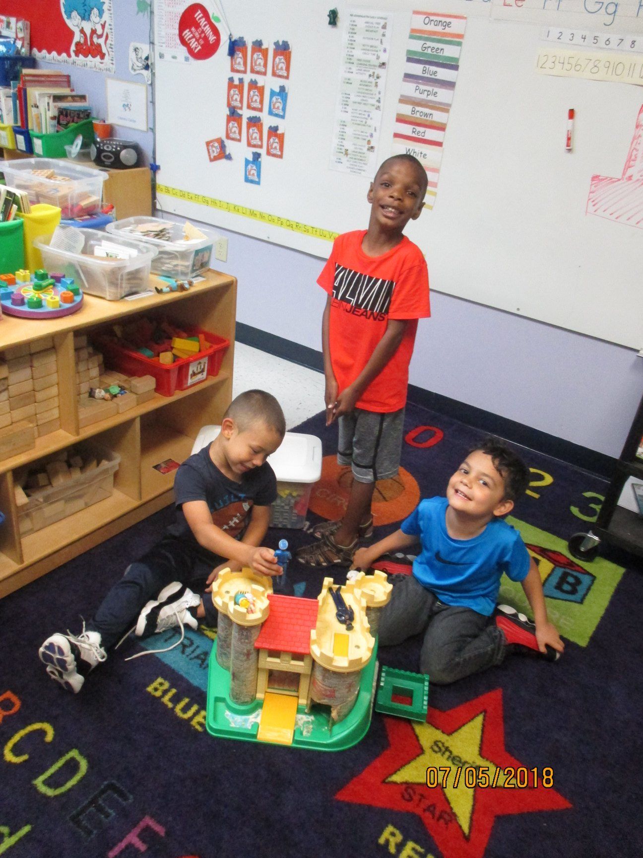 Three smiling children playing with a toy castle on a blue rug in a classroom.