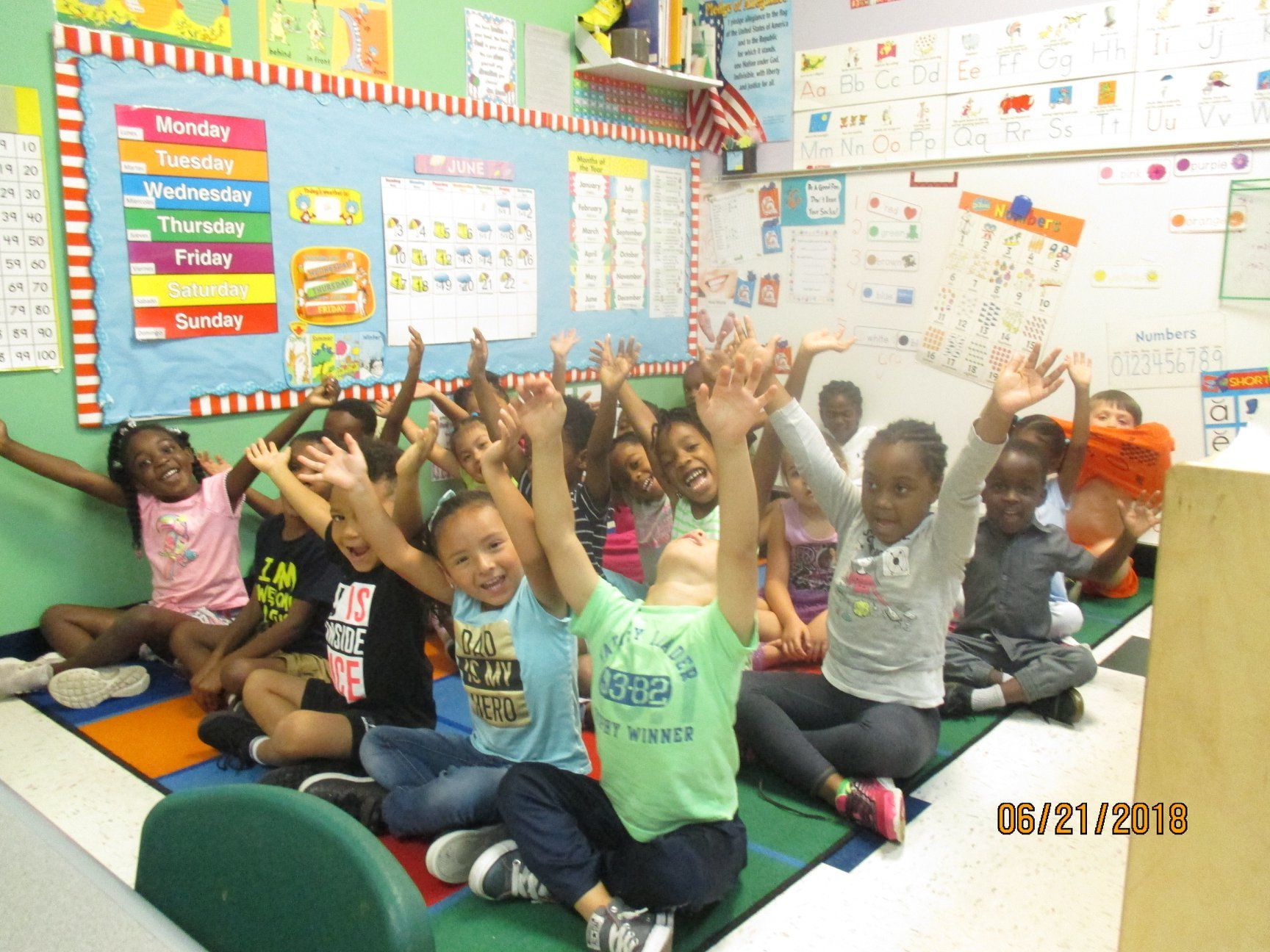 Children in classroom, arms raised, smiling. Colorful walls with calendar, charts.
