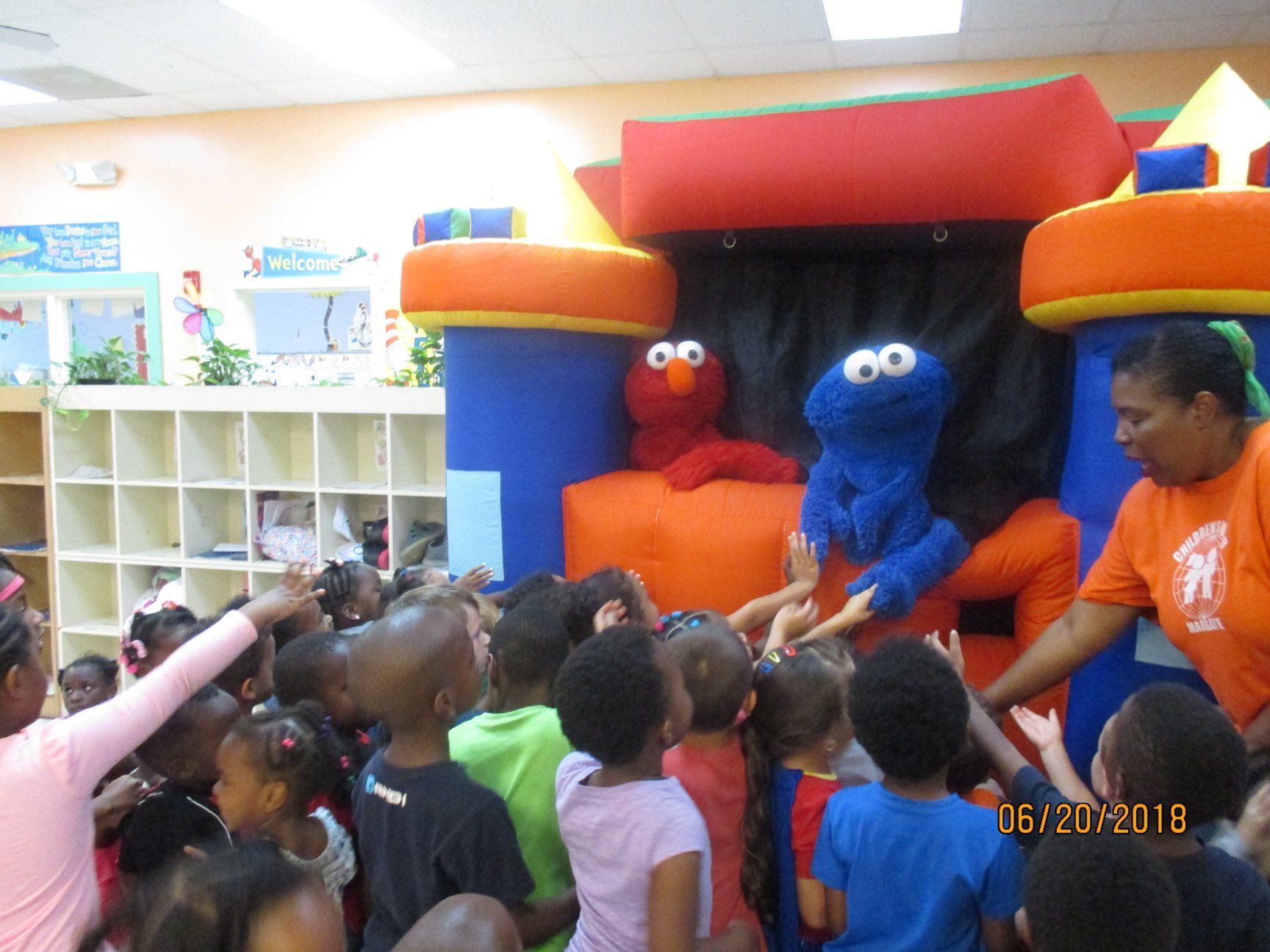 Children gather, reaching toward Elmo and Cookie Monster puppets, in front of an inflatable castle with a teacher.