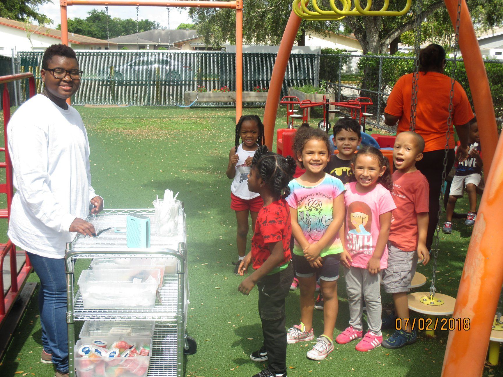 Teacher and children on a playground; teacher has a cart with supplies. Children are smiling.