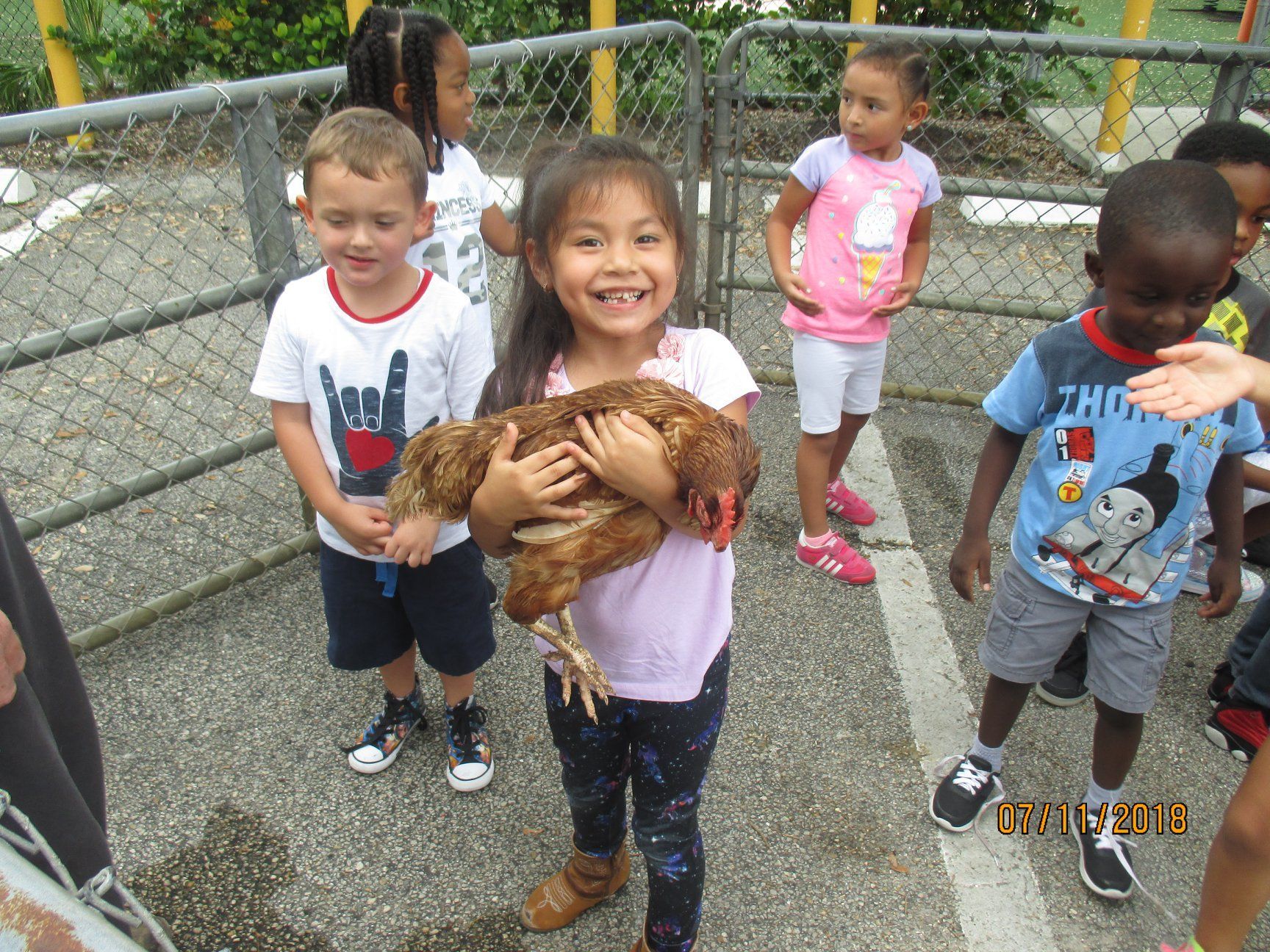 Girl holding a chicken, surrounded by smiling children in an outdoor enclosure.