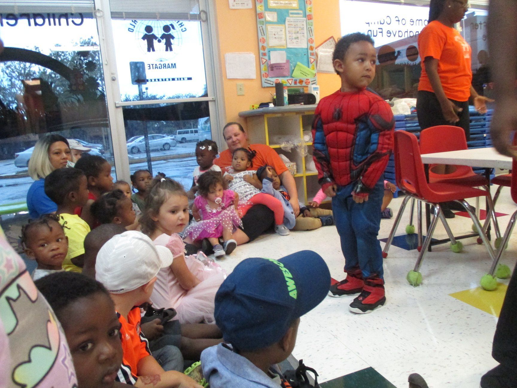 Children in costumes at a party; one boy in a Spider-Man suit stands as others sit, looking on.
