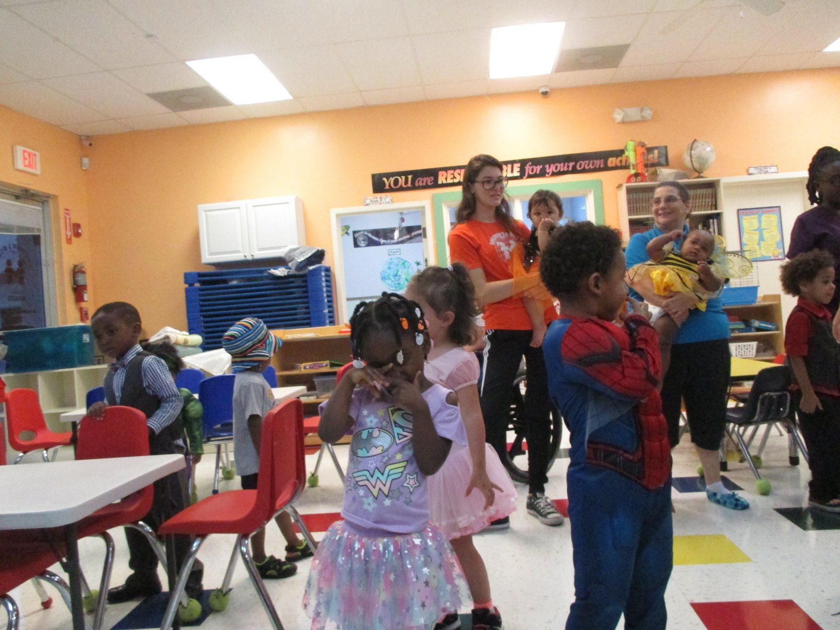 Children in costumes with teachers in a classroom. Orange and pink walls, red chairs, and white tables.