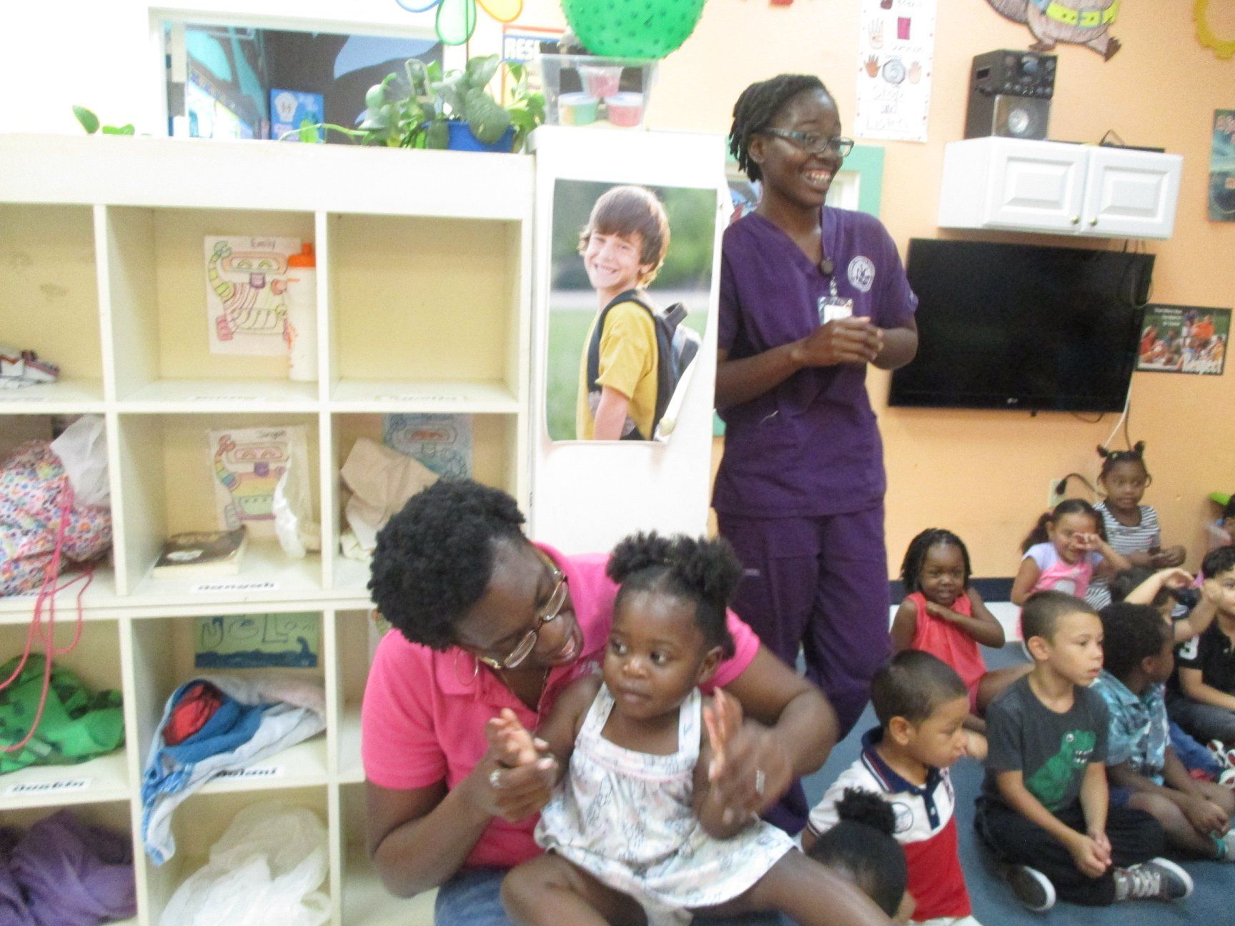 A woman in pink holds a child on her lap in a classroom, while another woman talks to a group of children.