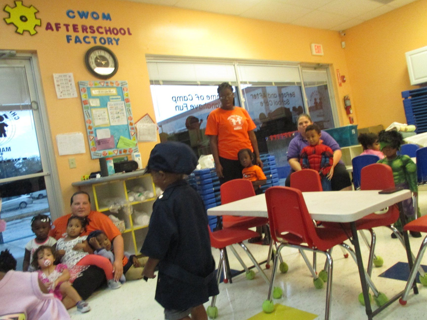 Children and adults in an after-school program. Several children are sitting or standing in the room, and some adults are present.
