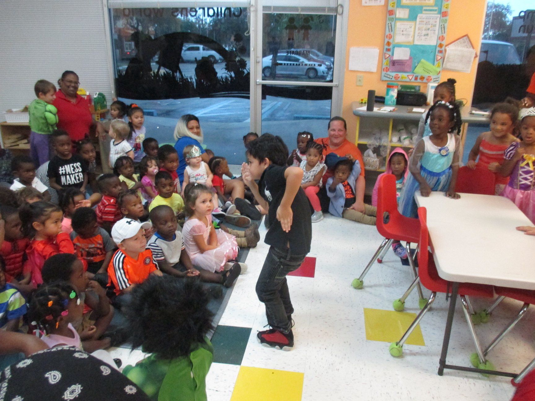 Children watching a performer in a classroom. Adults supervise. Colorful room with windows and tables.