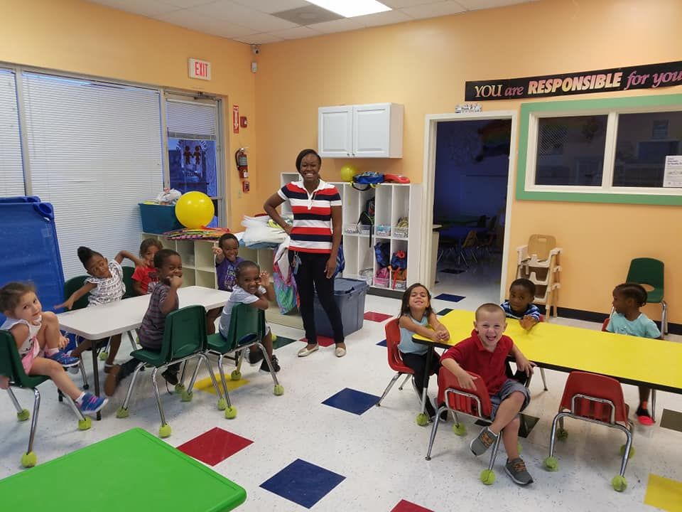 Teacher and diverse group of children in a classroom. They are sitting at tables, smiling. Brightly colored room.