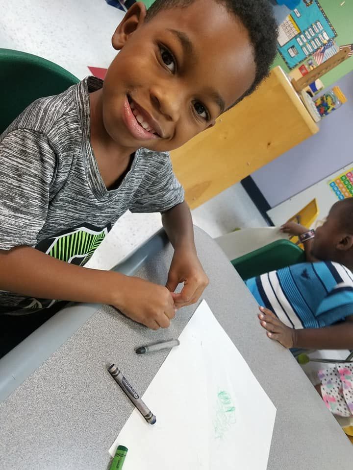Young boy smiles while drawing with crayons at a desk in a classroom.