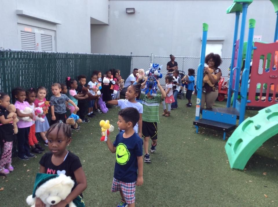 Children on a playground holding toys, being led by two adults, one holding a blue robot.