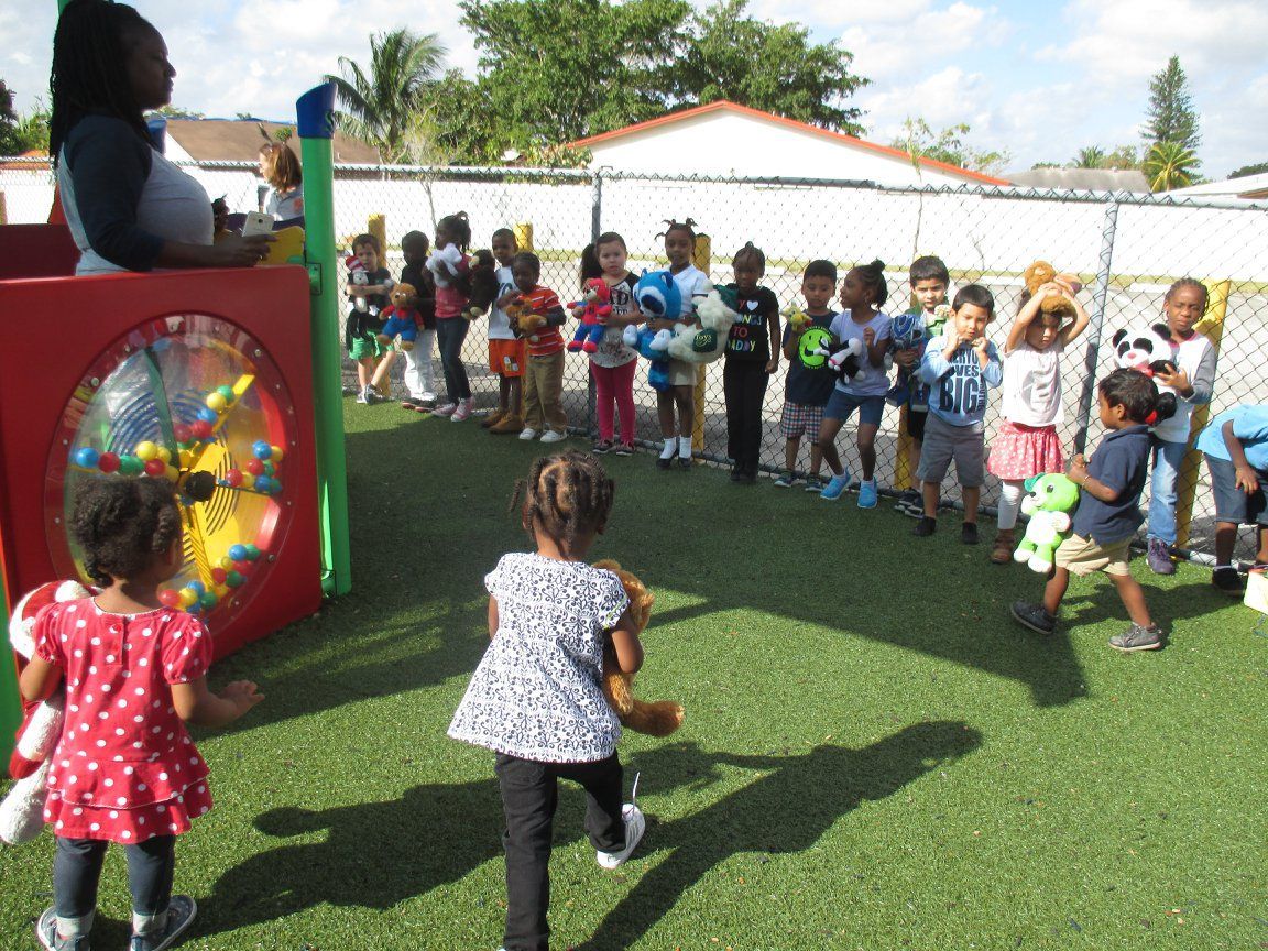 Children on a green lawn, playing a game with stuffed animals, watched by an adult. Sunny day.