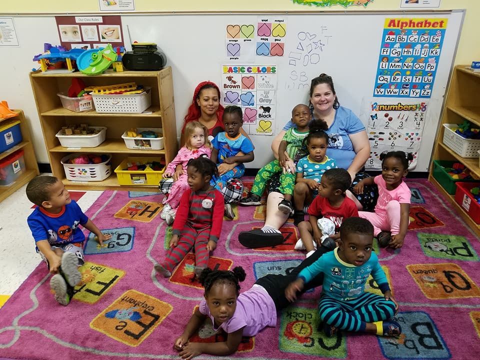Children and two teachers in a colorful classroom setting. Kids in pajamas are gathered on a rug.