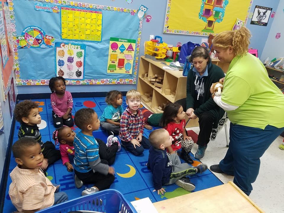 Children and teachers gather in a classroom, listening. Blue carpet. Teachers hold a small animal.