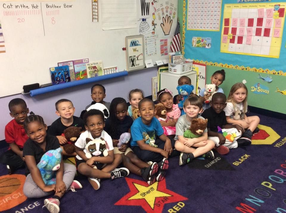 A group of children sitting on a colorful rug in a classroom, holding teddy bears.