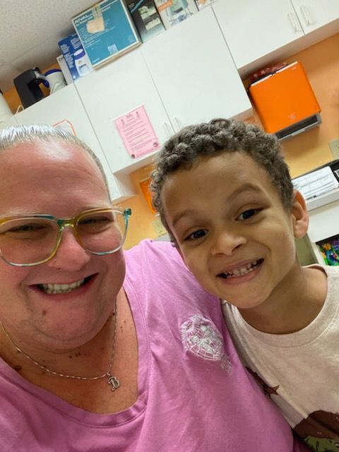 Woman and young boy smiling for a selfie in a brightly lit room. The woman wears glasses, a pink shirt, and the boy a white shirt.