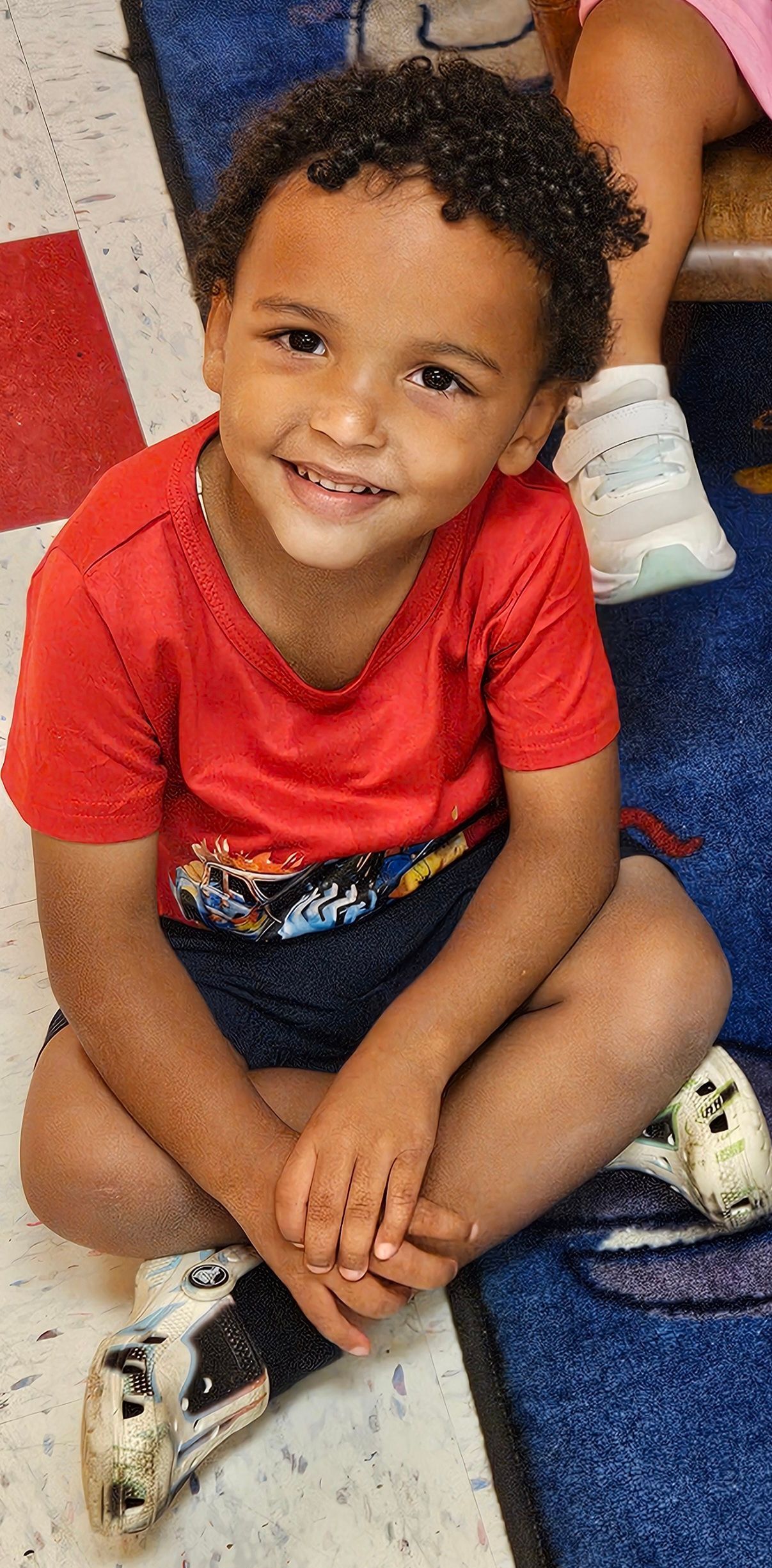 A smiling child with curly hair in a red shirt and shorts sitting on the floor.