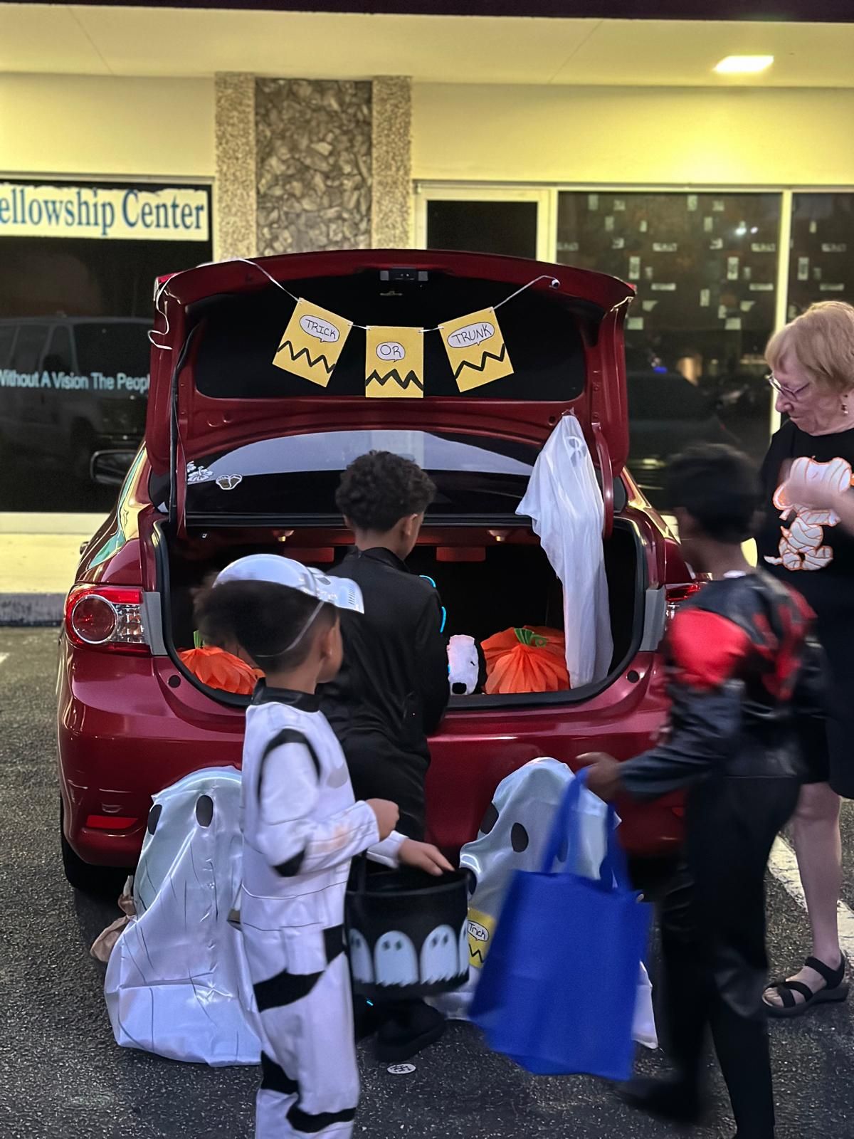 Children trick-or-treating at a trunk-or-treat event, dressed in costumes. Red car trunk decorated with Halloween garland.