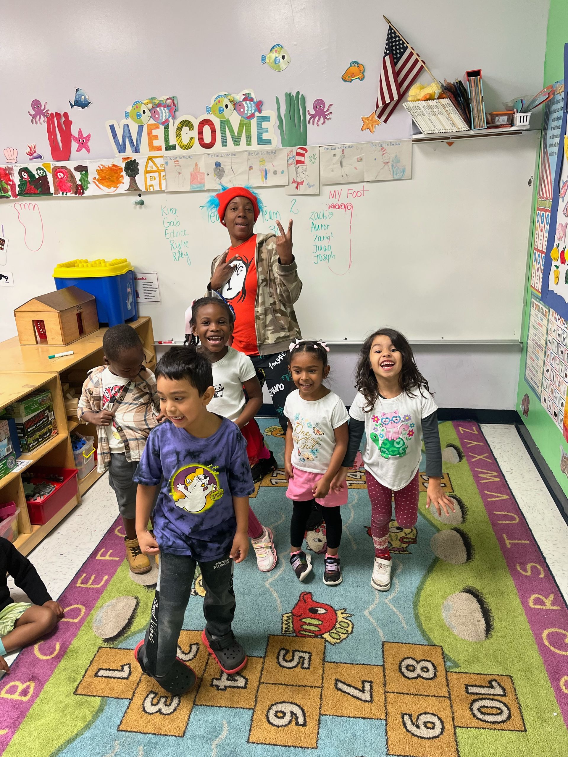 Children playing hopscotch in a classroom. Smiling, diverse kids stand on numbers, near a welcome sign.