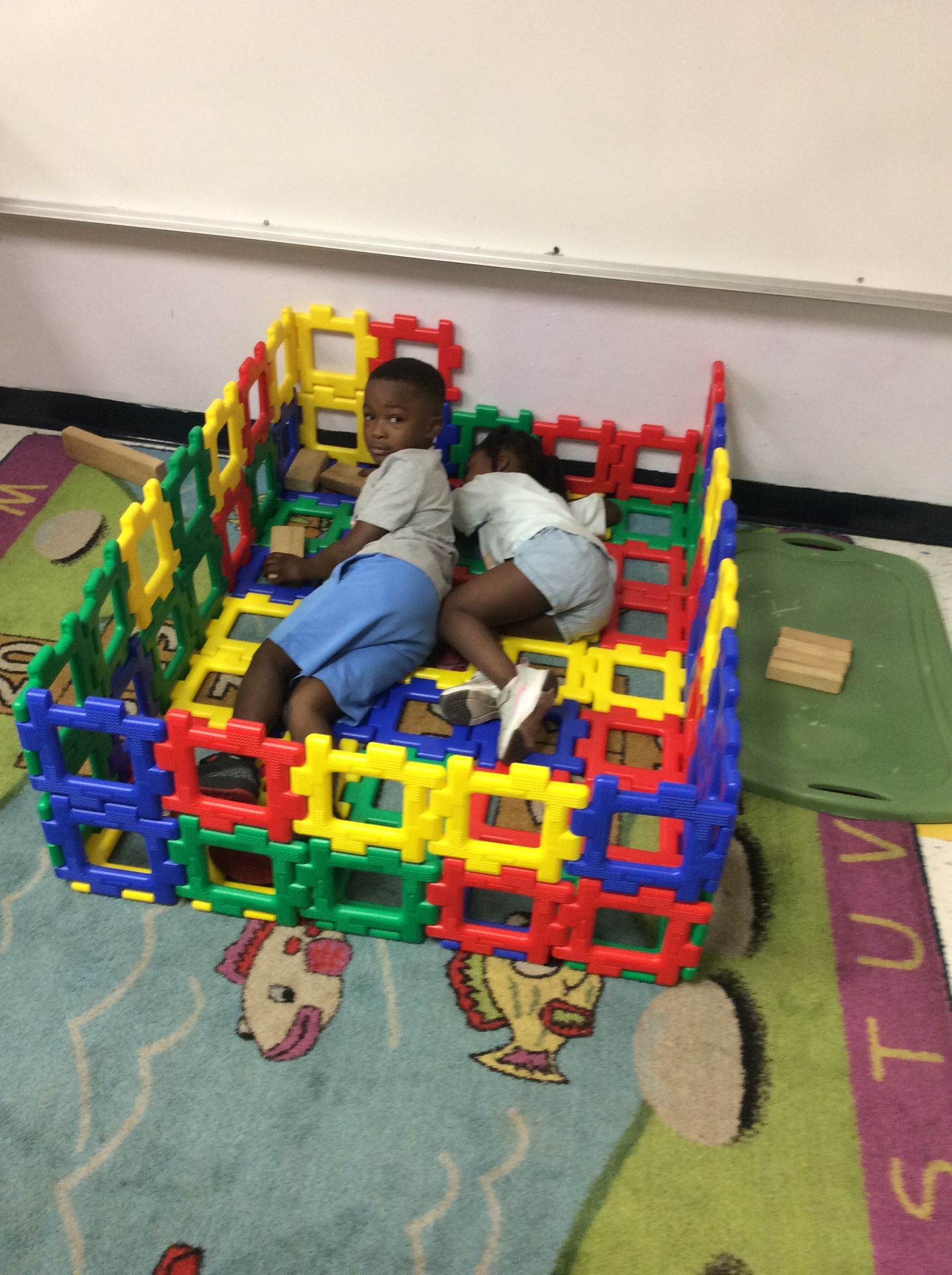 Two children inside a colorful block structure on a carpet in a room.
