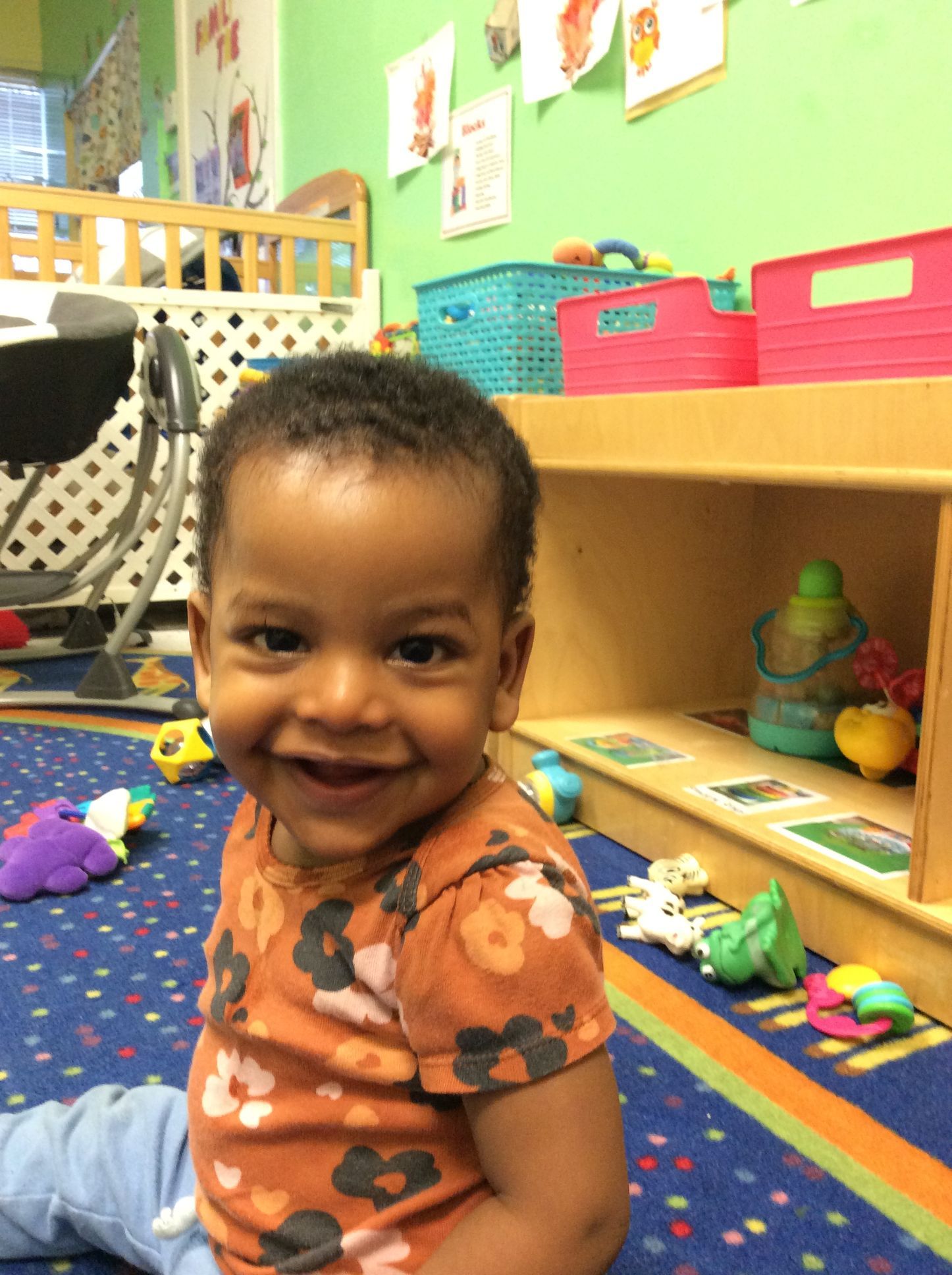 Smiling toddler in orange floral shirt sits on a blue carpet, toys scattered, green wall background.