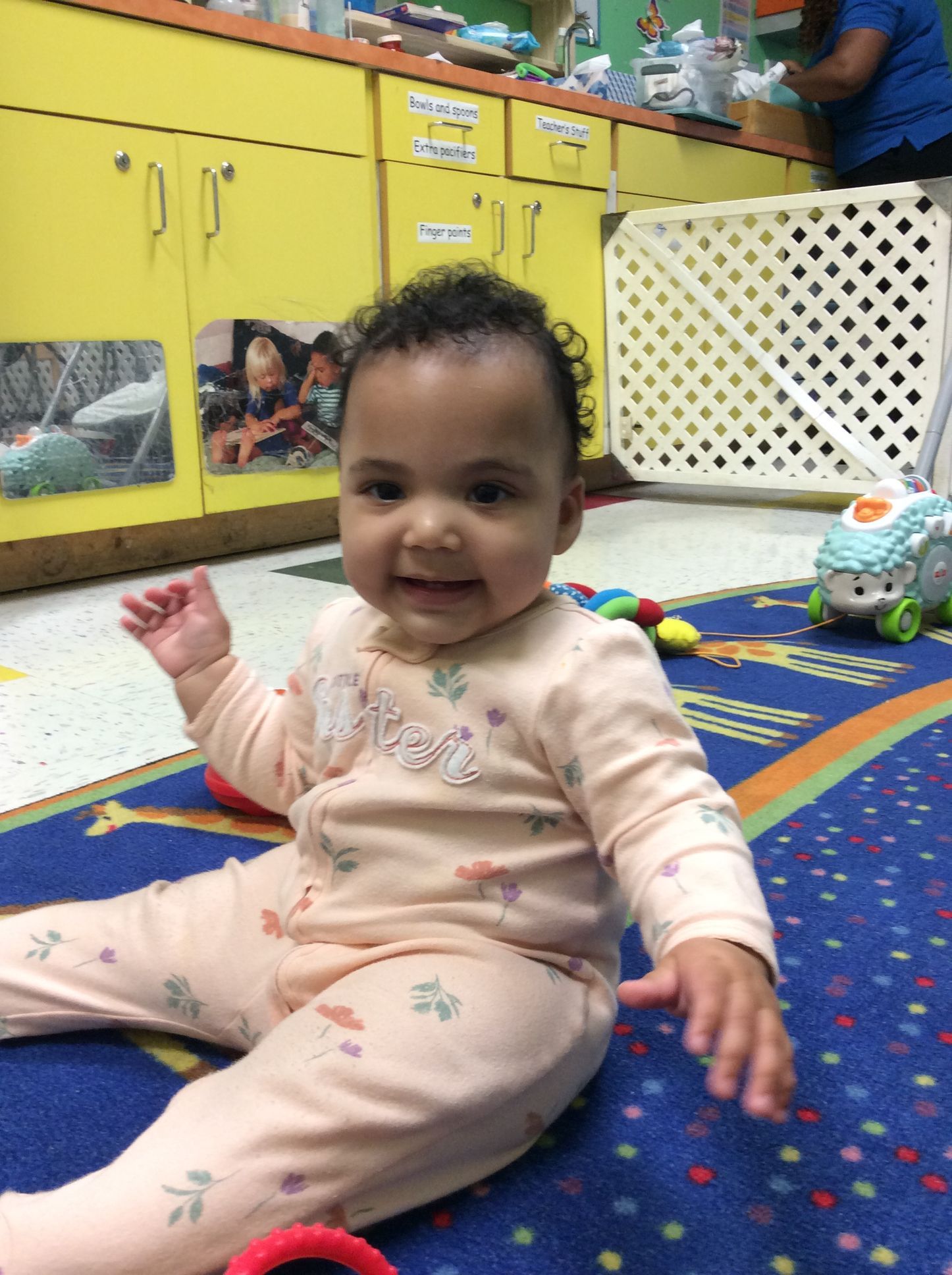 Smiling baby in peach onesie sitting on a blue patterned rug in a daycare setting.