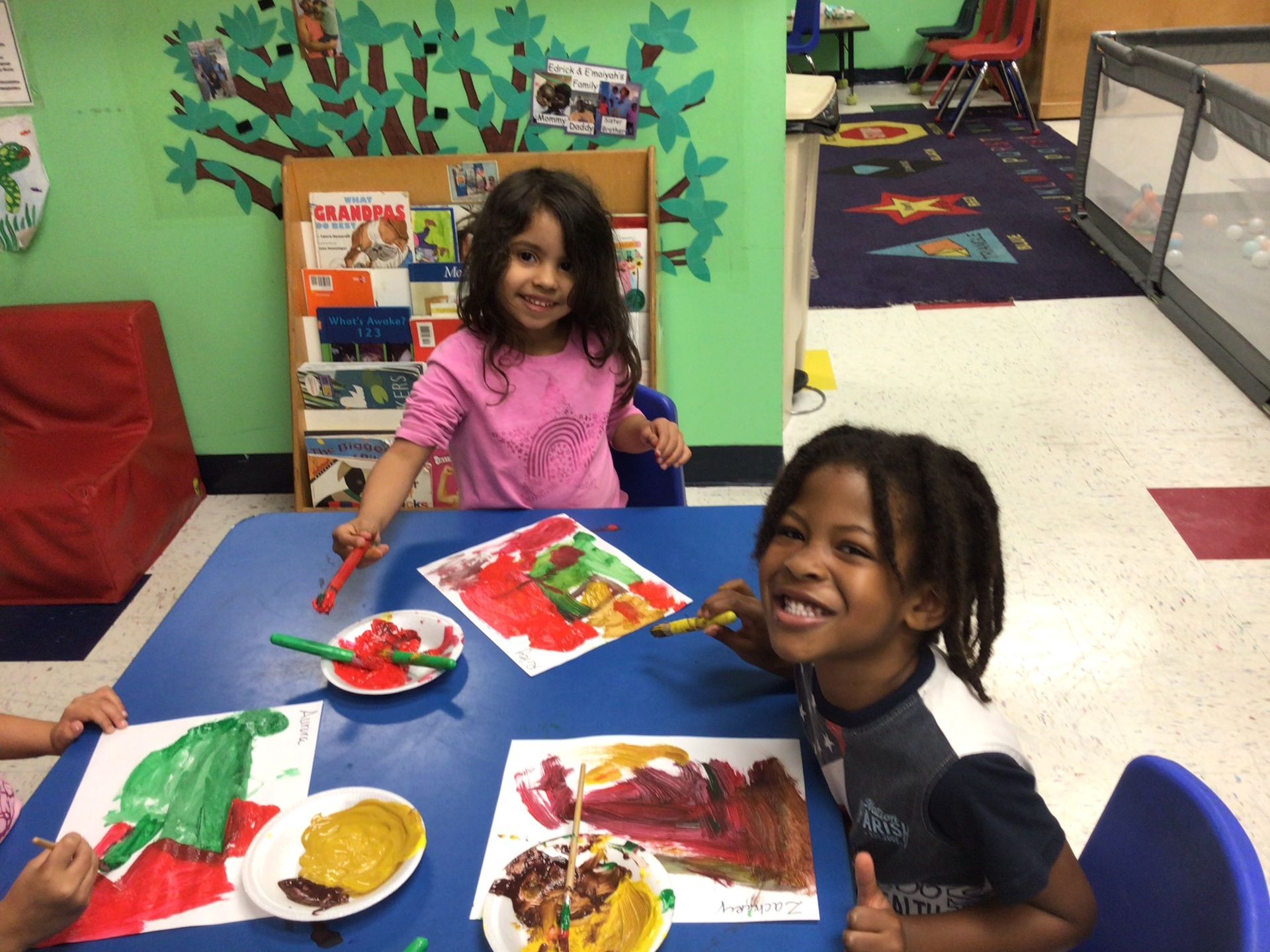 Two children smiling while painting at a table in a classroom; plates of paint and art on the table.
