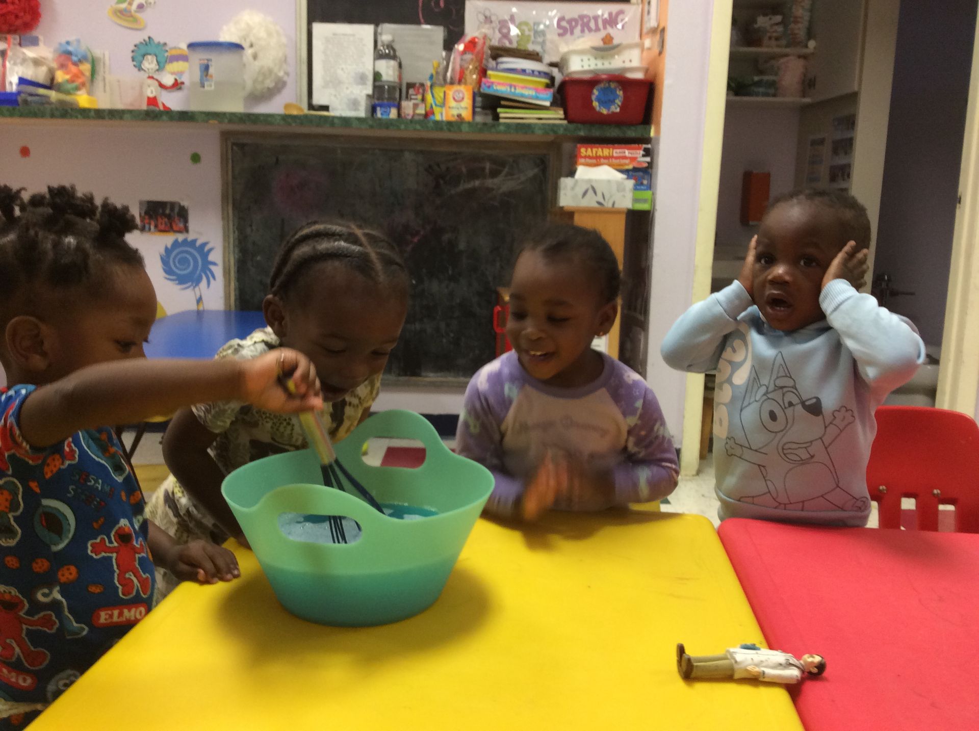 Four Black children at a table, playing with water and toy figures. One covers ears, the others react excitedly.