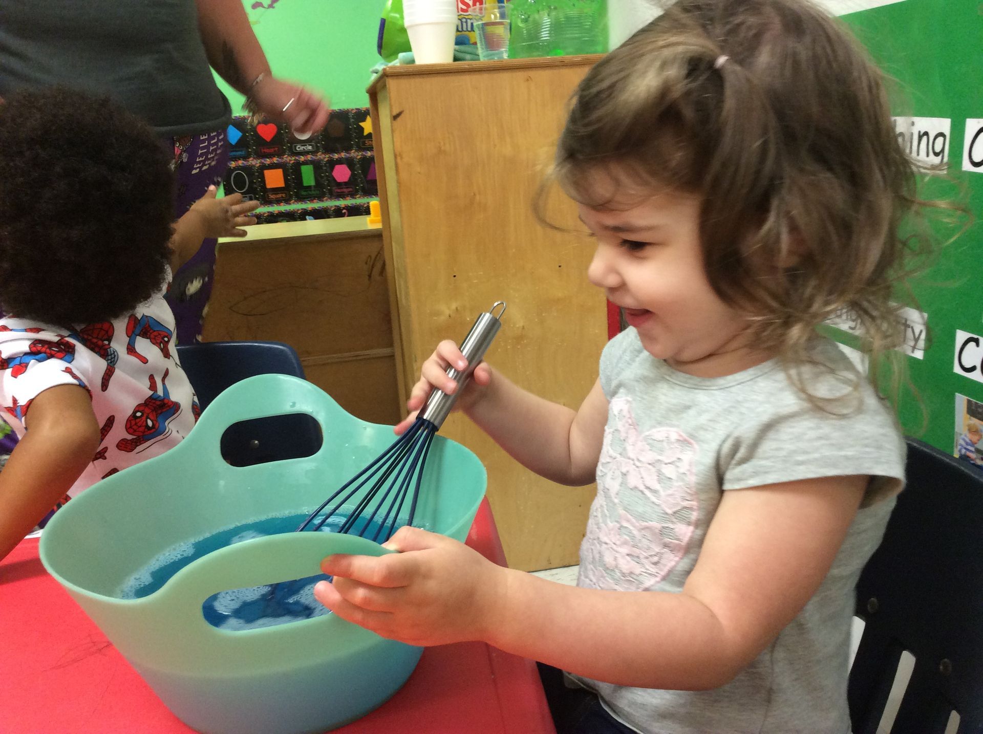 Girl stirring blue liquid in a bowl at a table with a whisk, another child nearby.