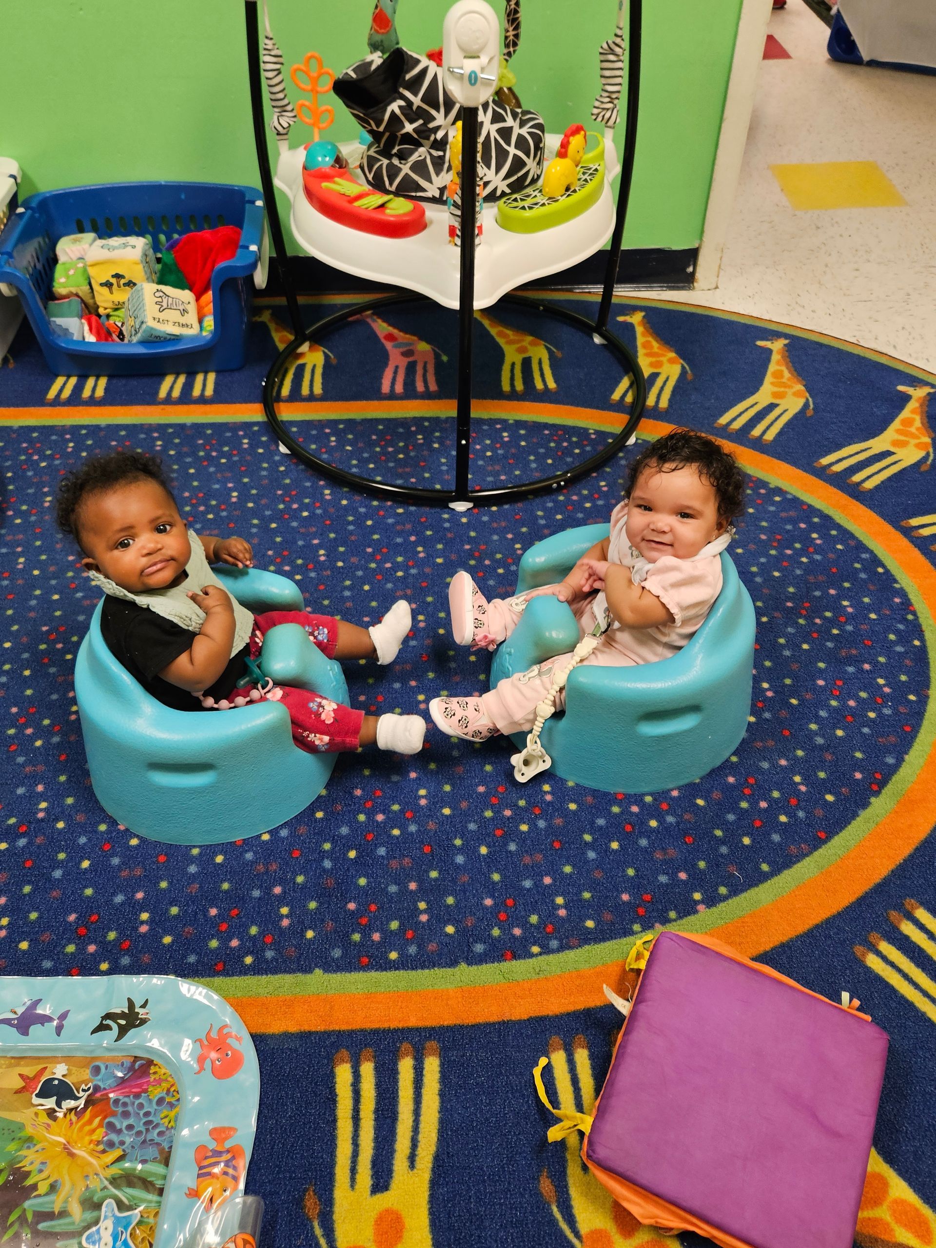Two babies sitting in blue chairs on a patterned rug, looking at the camera.